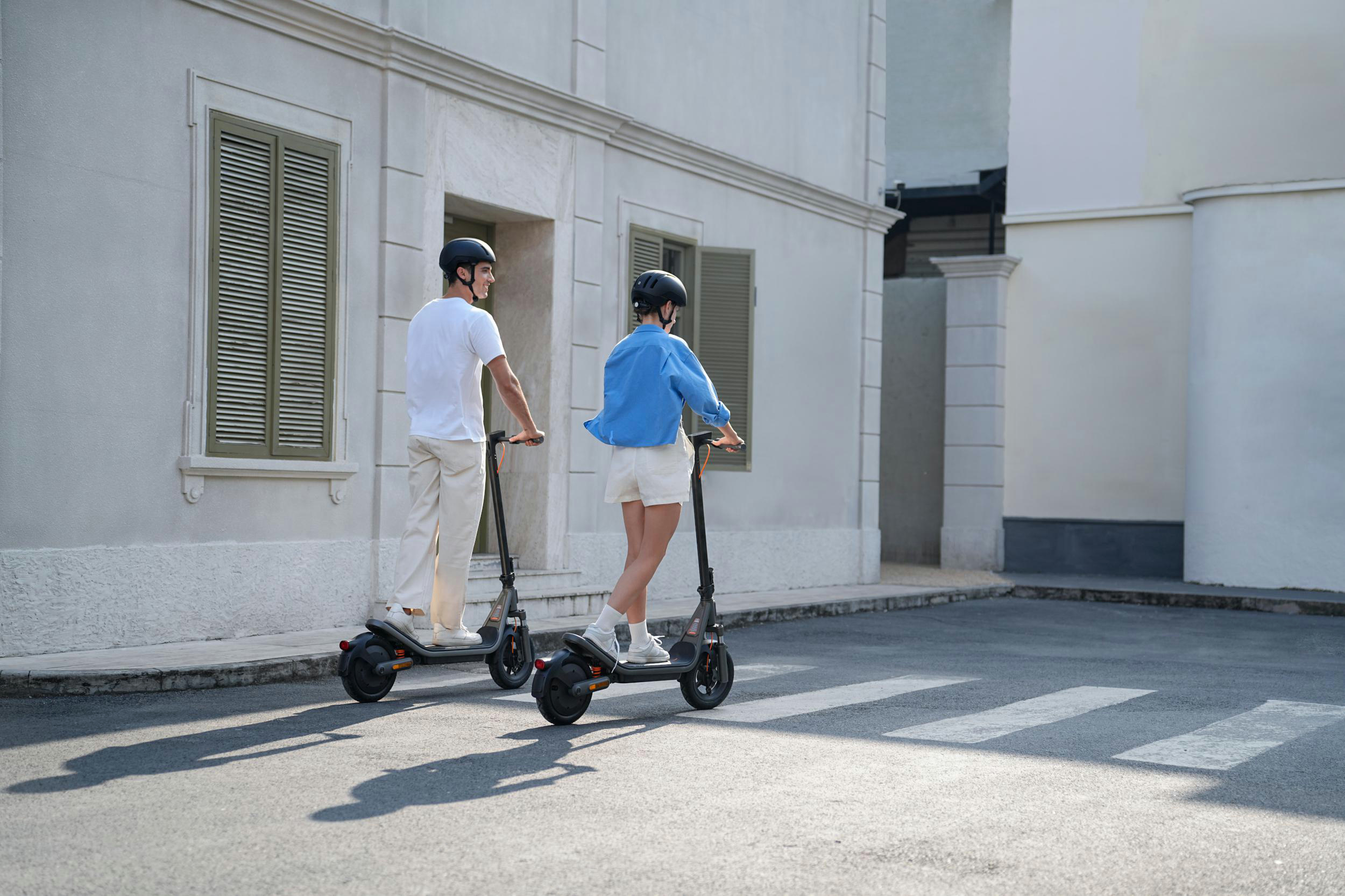 Zwei Personen fahren Roller auf einer Straße vor einem Gebäude.