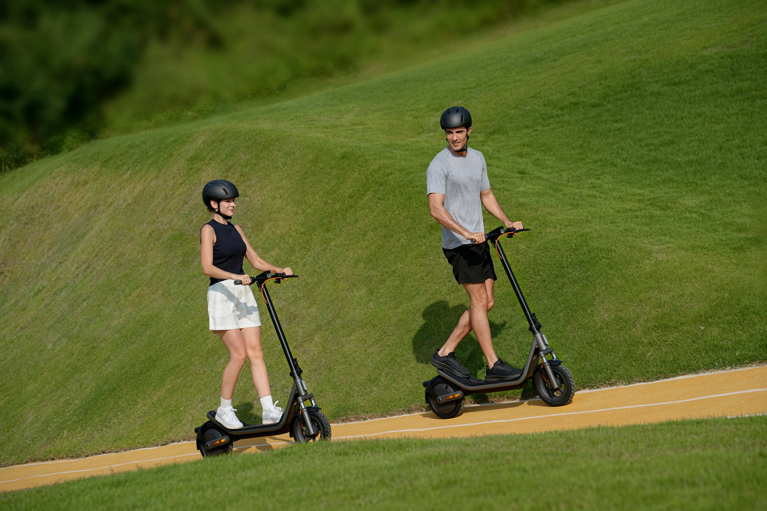 Mann und Frau fahren E-Scooter auf einem gelben Weg in einer grünen Landschaft.