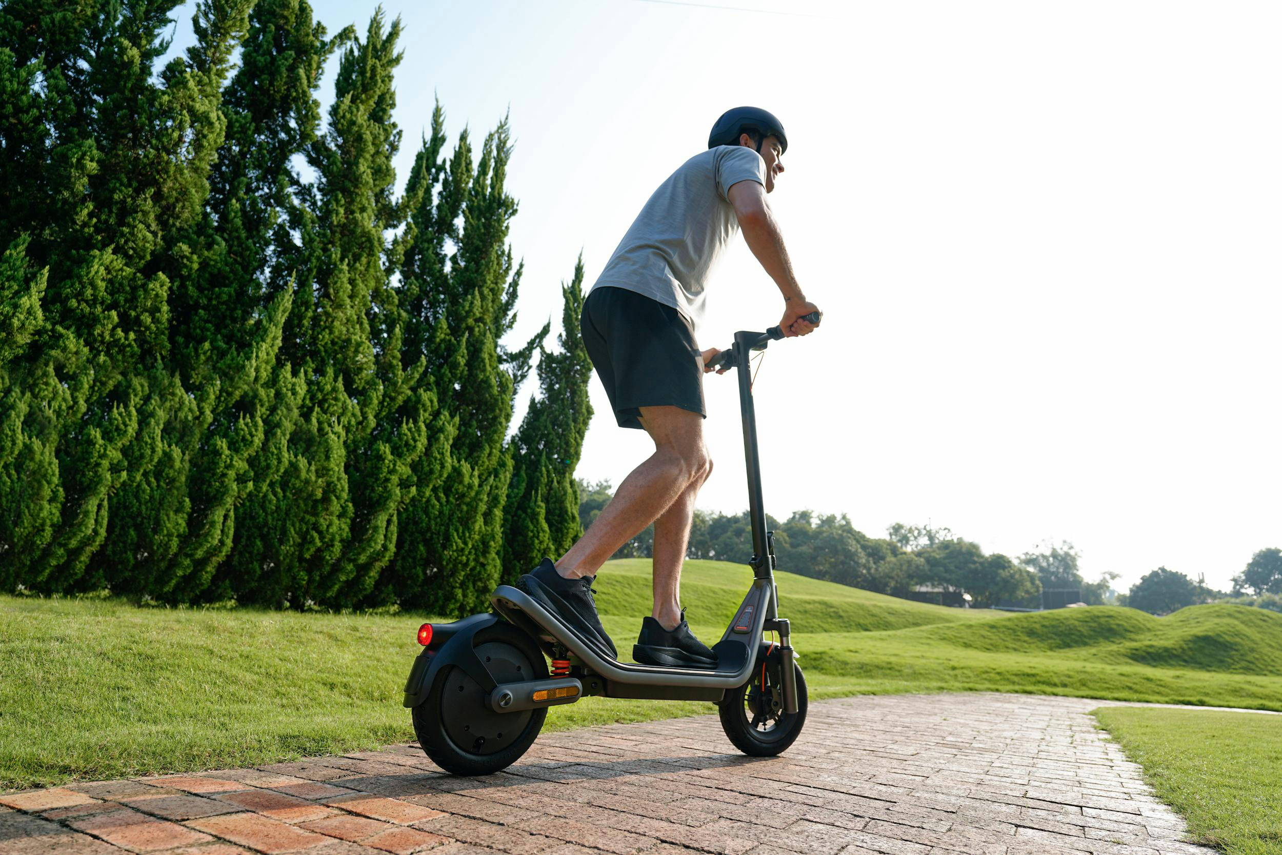 Mann auf E-Scooter fährt auf einem gepflasterten Weg neben Bäumen.
