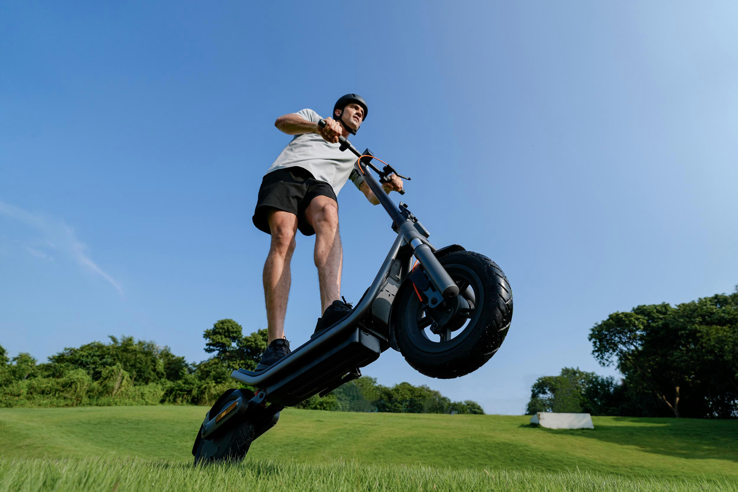 Mann auf Elektroroller fährt im Gras, blauer Himmel.