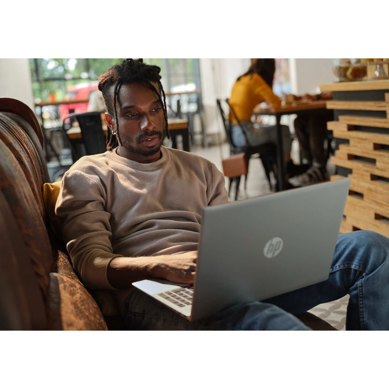 Man met dreadlocks zit op bank laptop te gebruiken. Mensen op achtergrond in café.