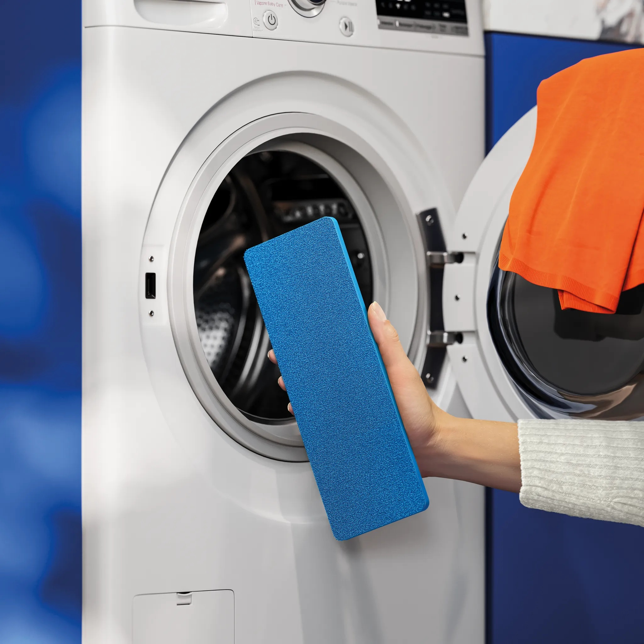 Ręka trzyma niebieską gąbkę przed otwartą pralką z pomarańczowymi ubraniami.
