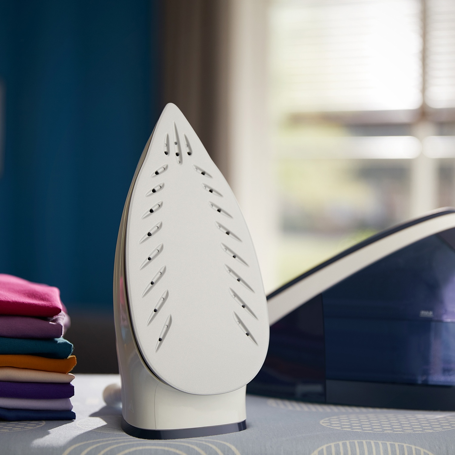 An iron rests next to a stack of colorful clothes on an ironing board.