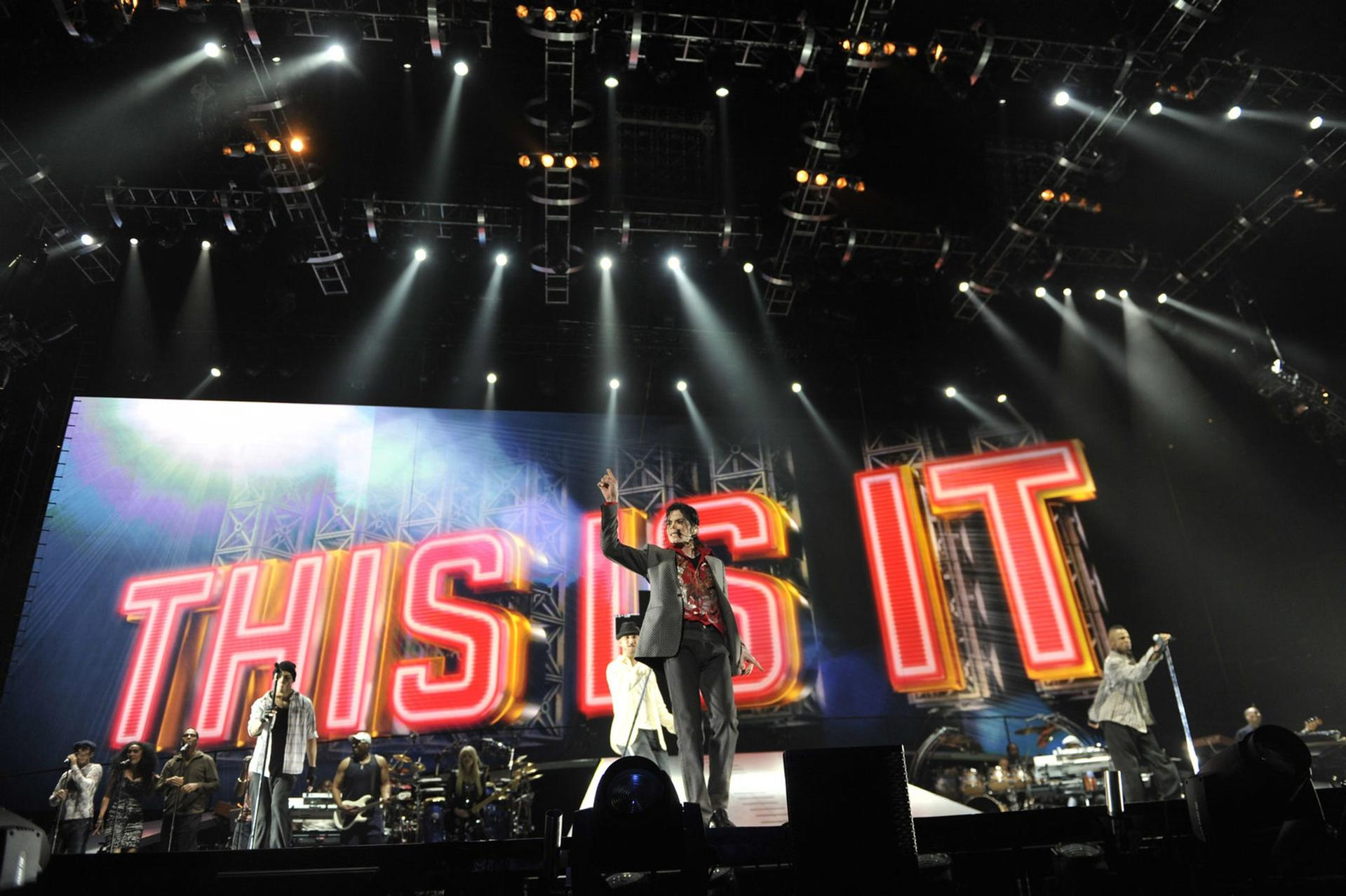 Michael Jackson und Band auf der Bühne vor großem "THIS IS IT"-Banner, beleuchtet von Scheinwerfern.
