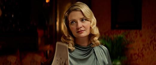 Eine Frau mit blonden Locken und einem grauen Kleid blickt nach rechts.