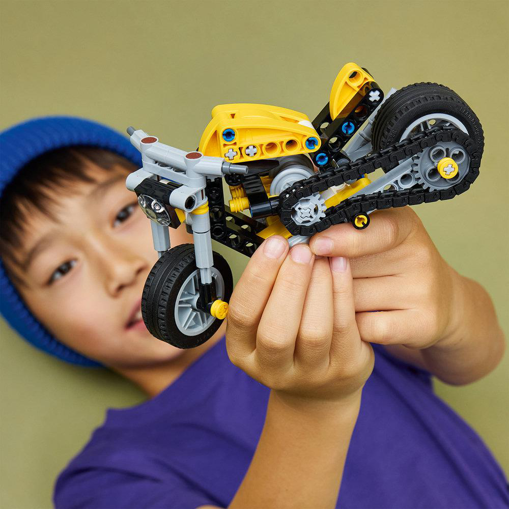 Ein Junge mit blauer Mütze hält ein gelb-graues Lego-Motorrad vor einem grünen Hintergrund.