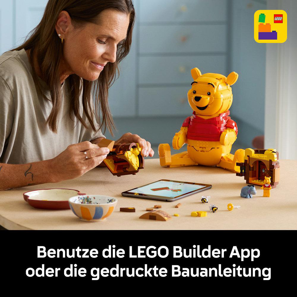 Eine Frau baut mit LEGOs, Winnie Puuh sitzt daneben. Ein Tablet zeigt eine Anleitung.