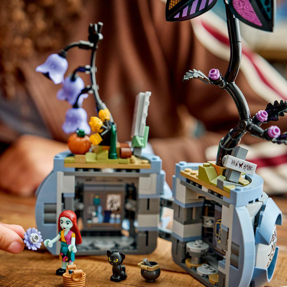 Sally Figur hält eine Blume vor Lego-Bauten mit lila Blumen und Kürbis.