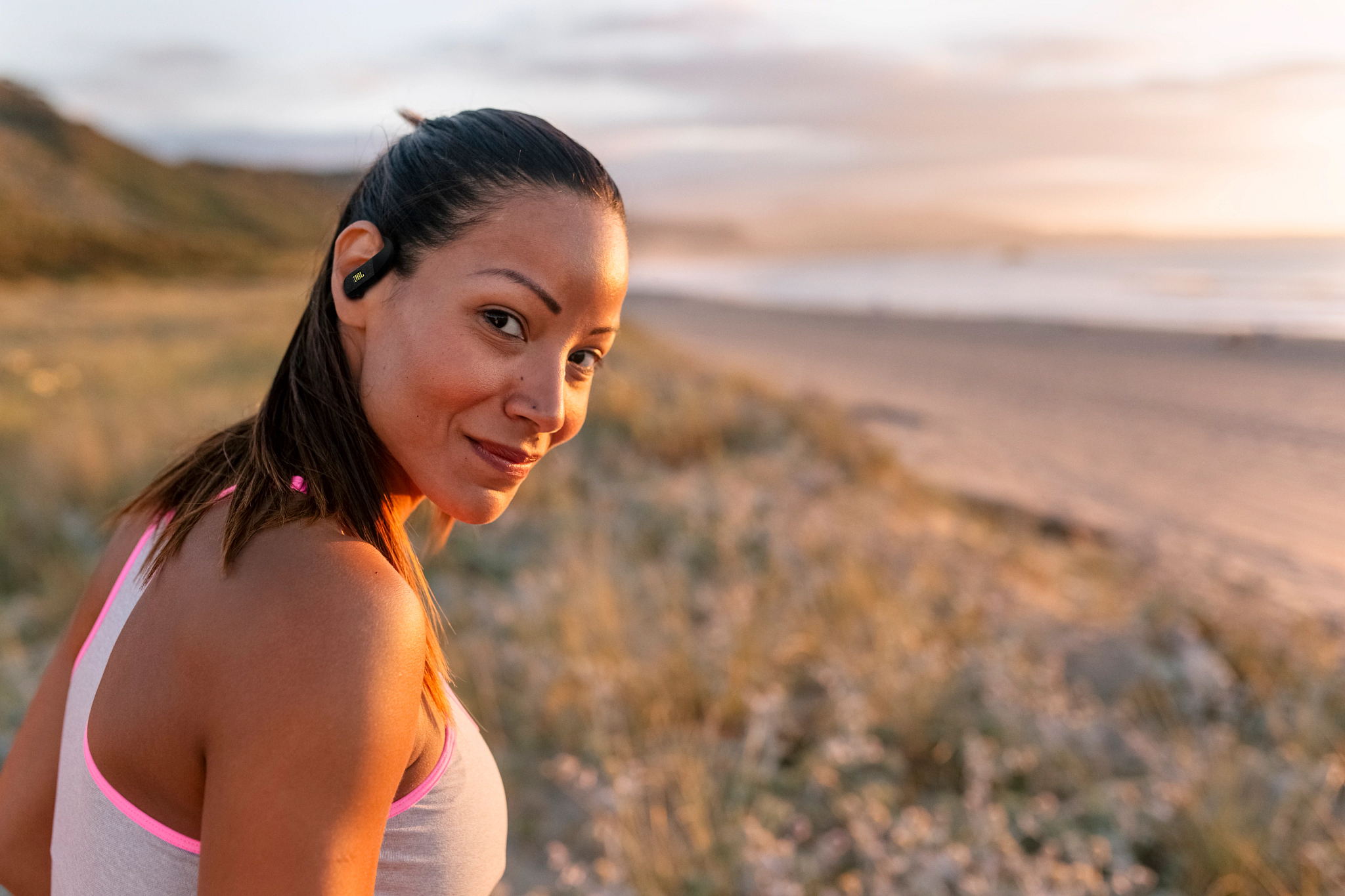 Frau mit Kopfhörern am Strand bei Sonnenuntergang, lächelt.