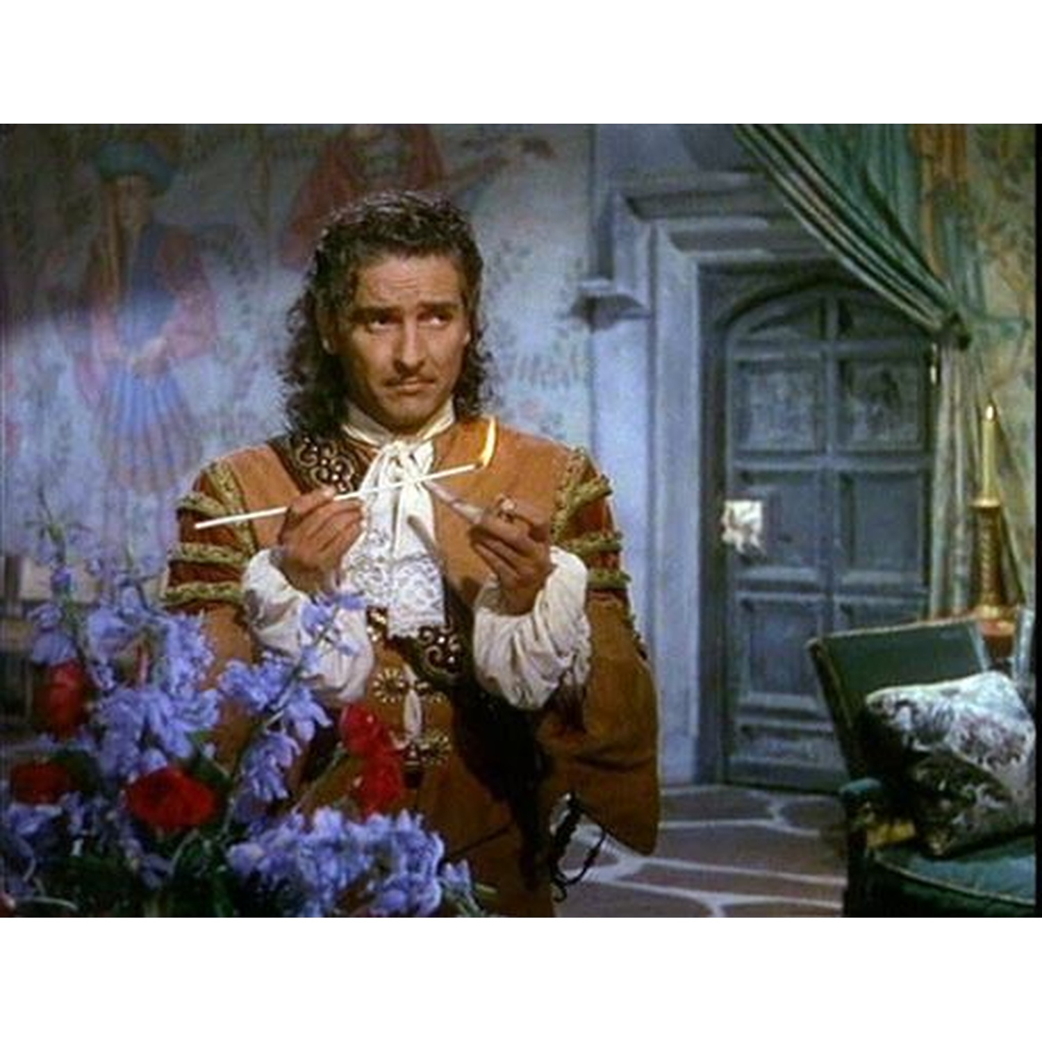 Man in historische kledij houdt rietstengel bij bloemen en kaars.