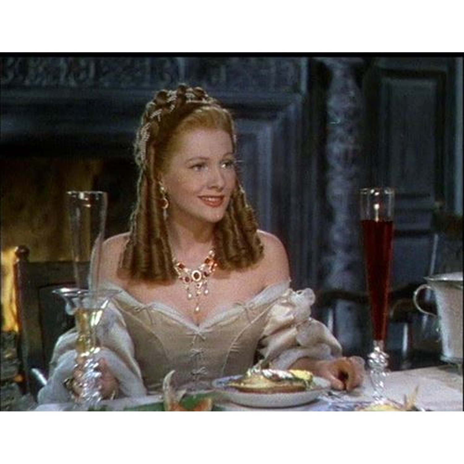 Een vrouw in een crèmekleurige jurk met sieraden zit aan tafel.