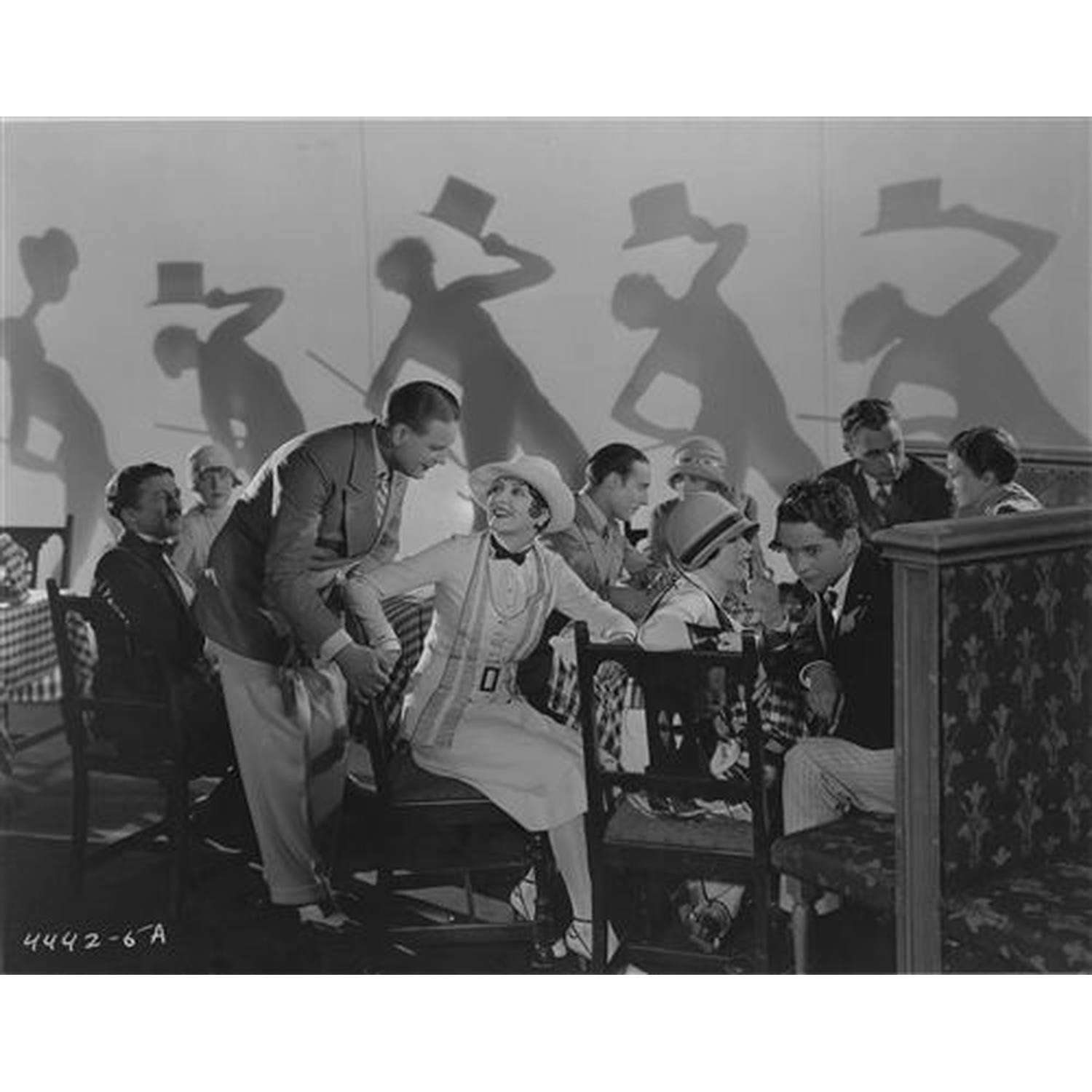 Un homme aide une femme au chapeau et à la robe. Des ombres de danseurs derrière.