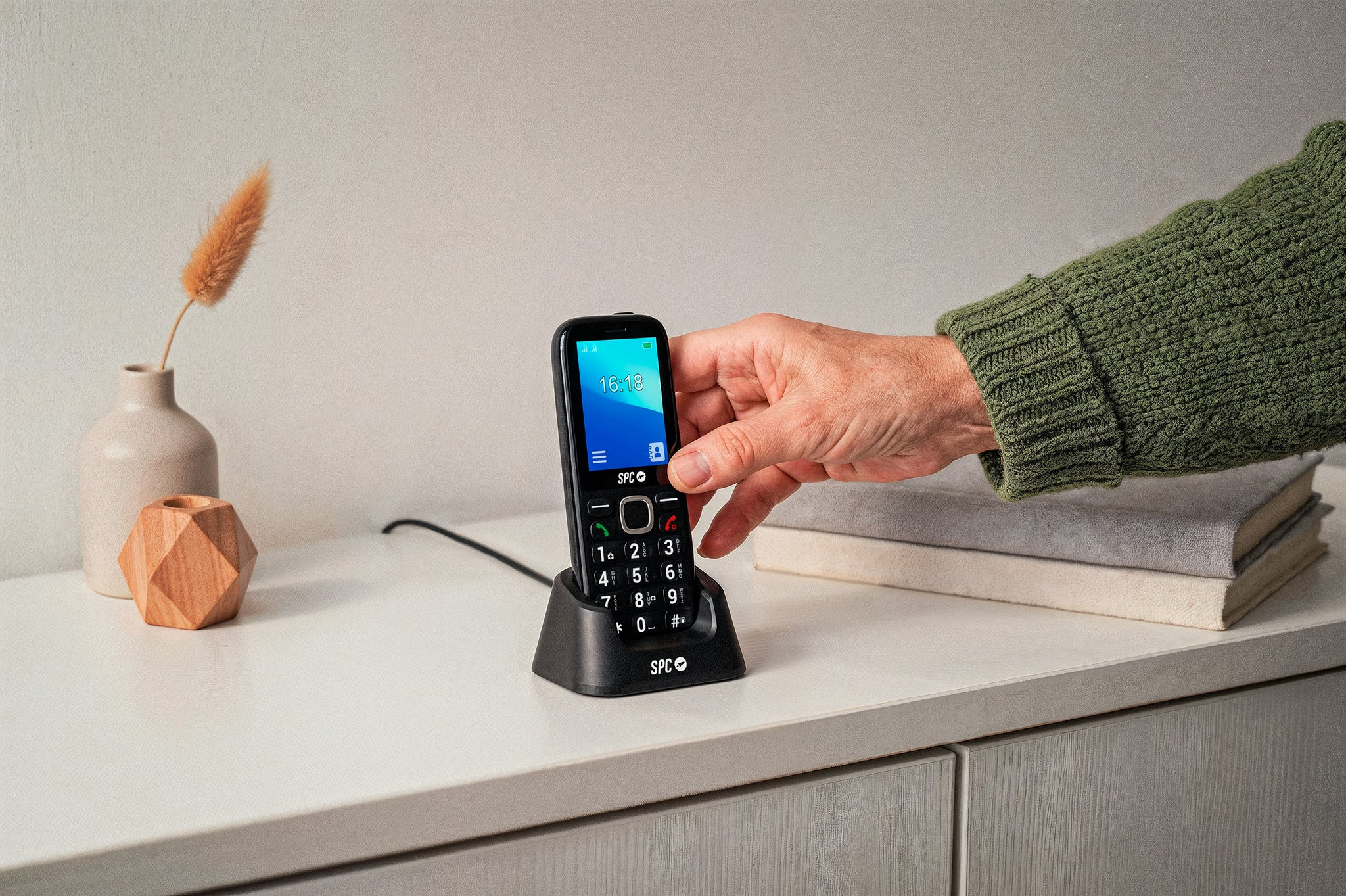 Mano coloca un teléfono SPC negro en una base de carga sobre una mesa blanca.