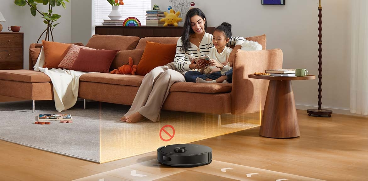 Robot sprzątający z czerwonym znakiem zakazu zbliża się do sofy, na której siedzą kobieta i dziecko.
