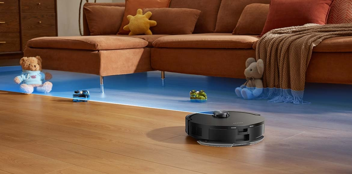 Czarny robot sprzątający czyści drewnianą podłogę w pobliżu brązowej sofy z zabawkami.