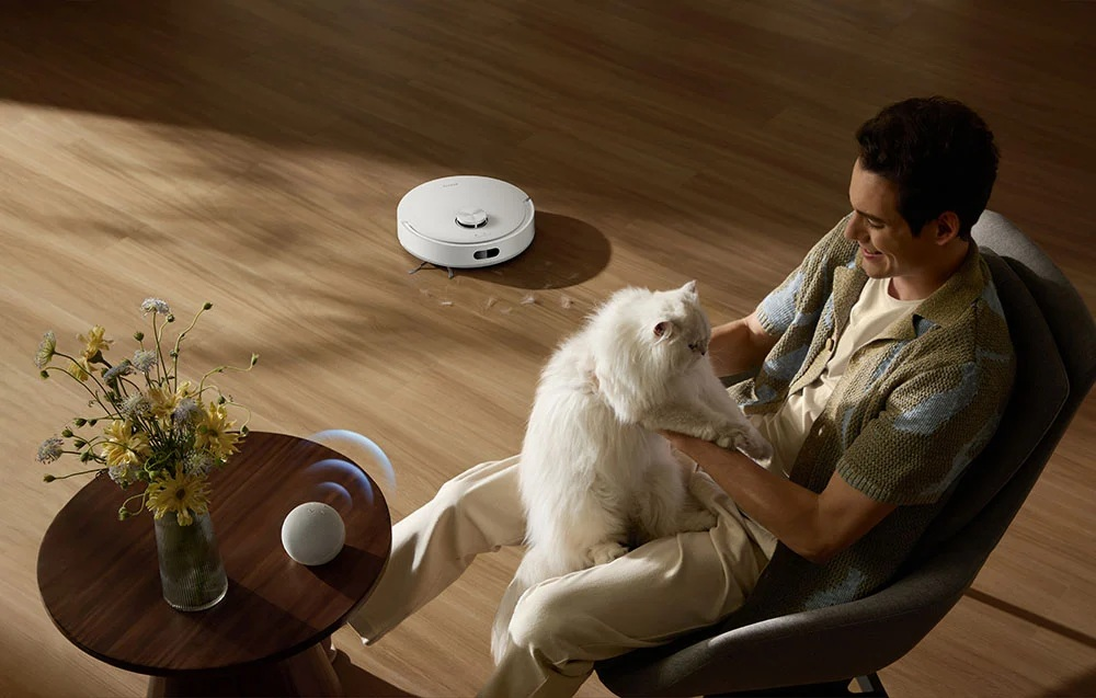 Mężczyzna siedzi z kotem, robot sprzątający czyści podłogę, kwiaty na stole.