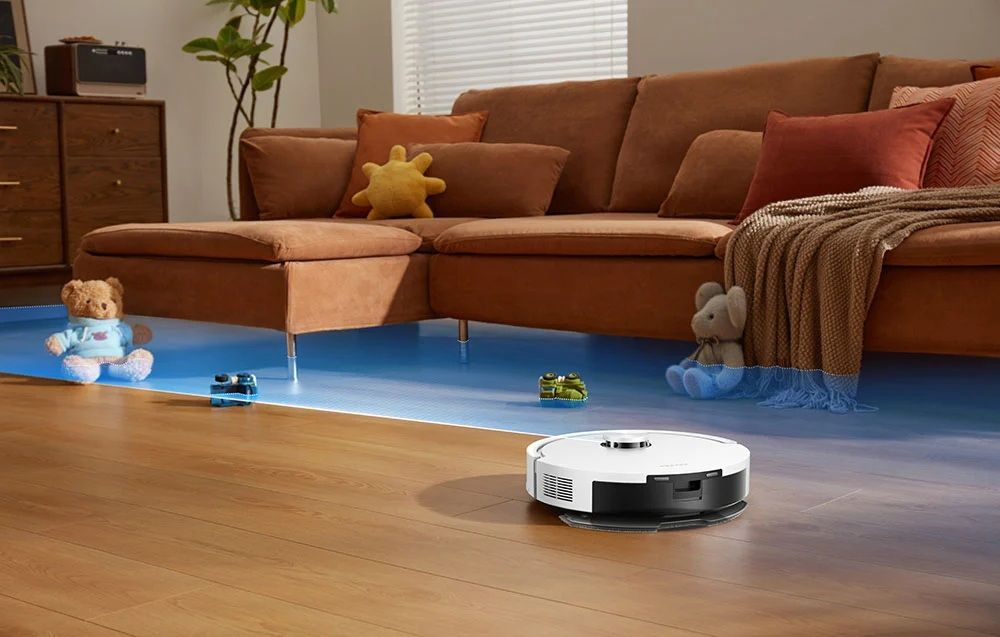 Biały robot sprzątający czyści drewnianą podłogę przed brązową sofą z zabawkami.