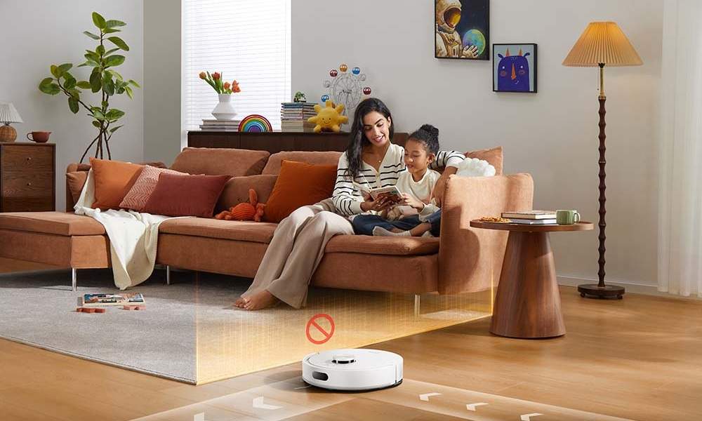 Odkurzacz automatyczny czyści dywan, podczas gdy matka i dziecko czytają na kanapie.