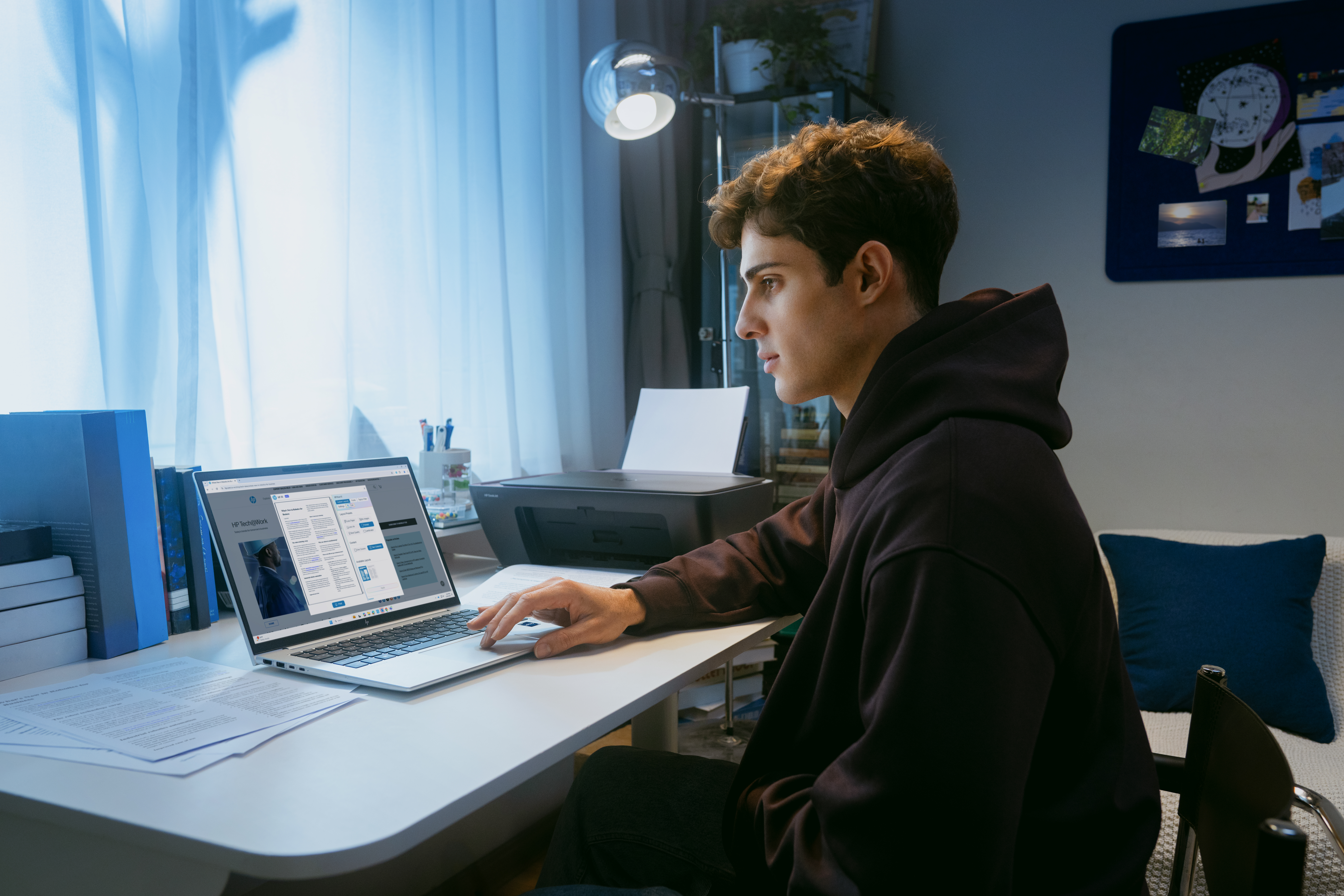 Ein junger Mann sitzt an einem Schreibtisch und arbeitet an seinem Laptop. Ein Drucker steht daneben.