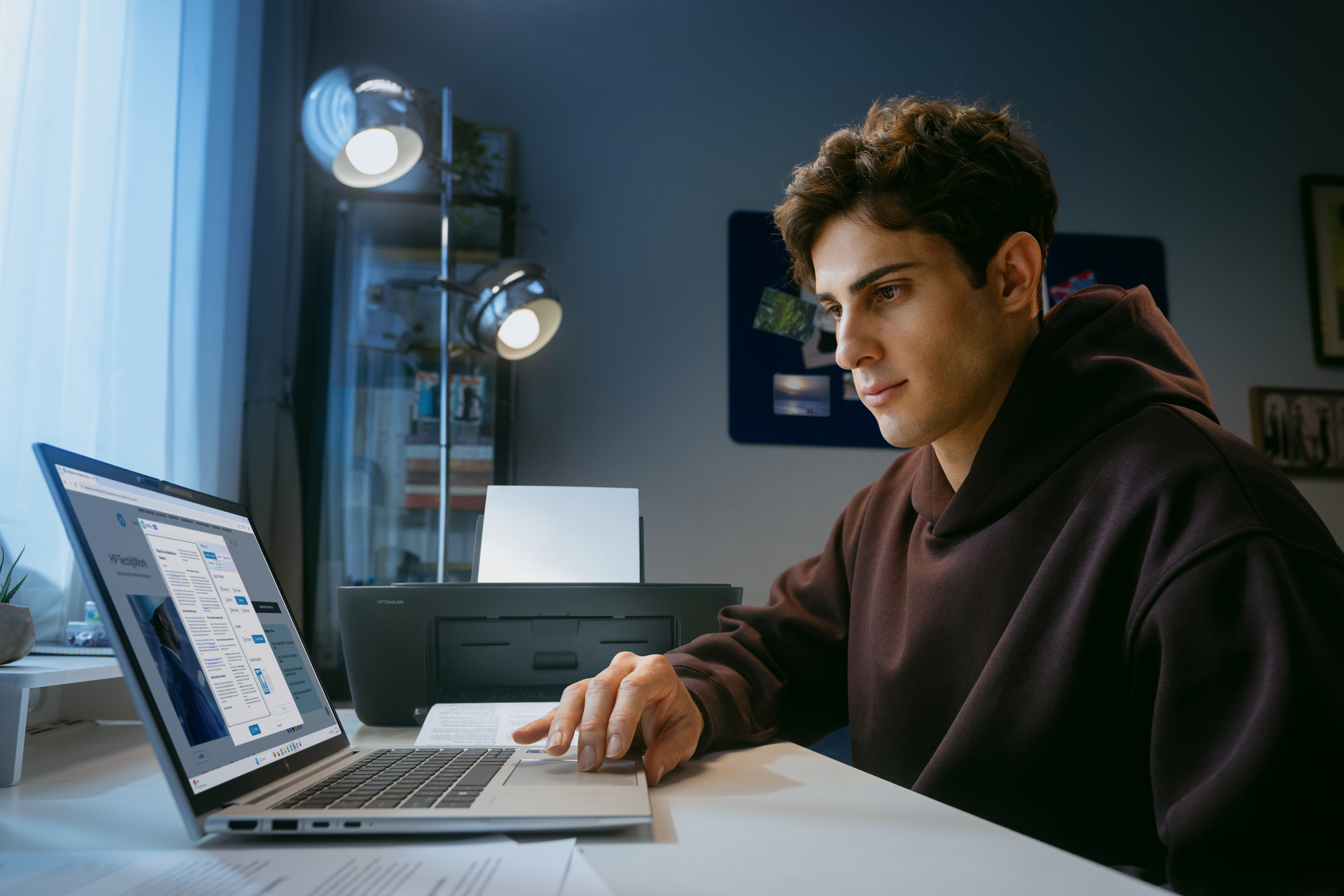 Ein Mann mit Kapuzenpullover arbeitet am Laptop, Drucker im Hintergrund.