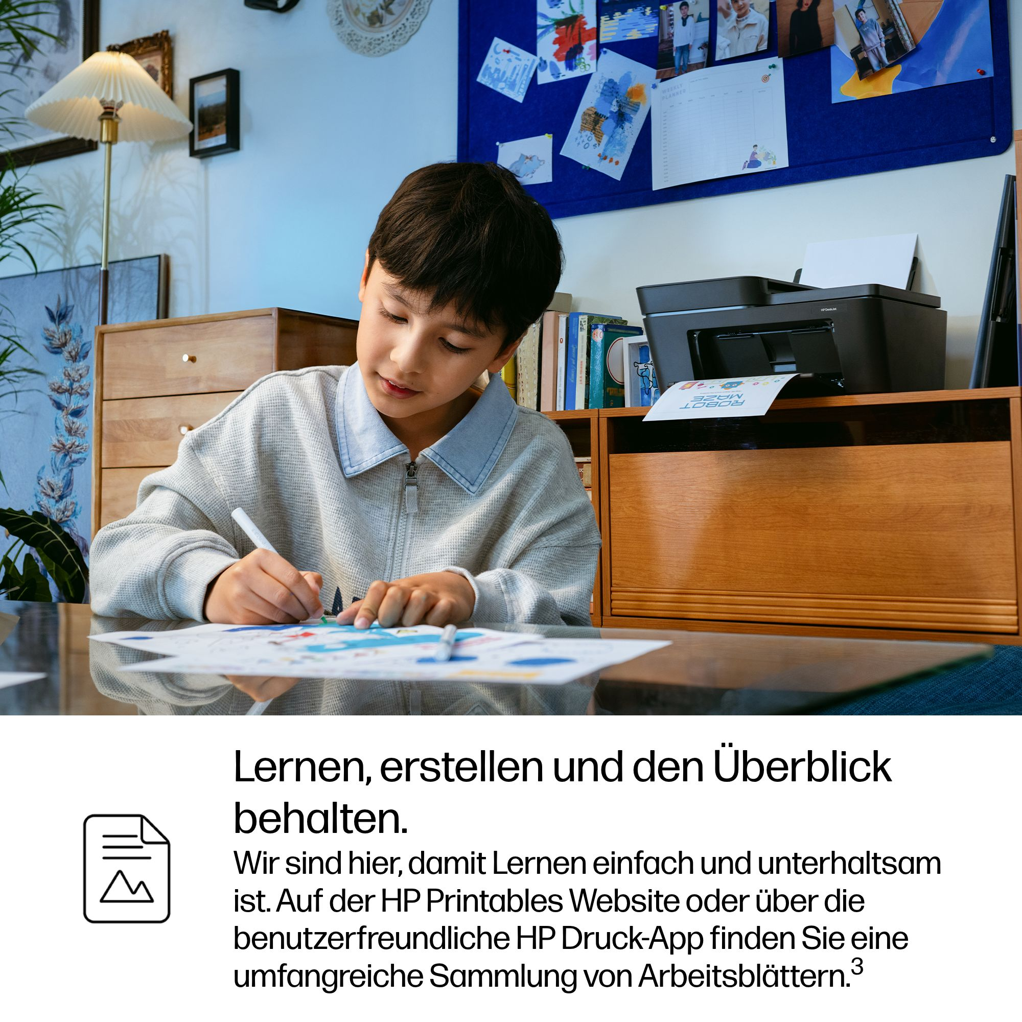 Junge zeichnet am Tisch, Drucker und Pinnwand im Hintergrund.