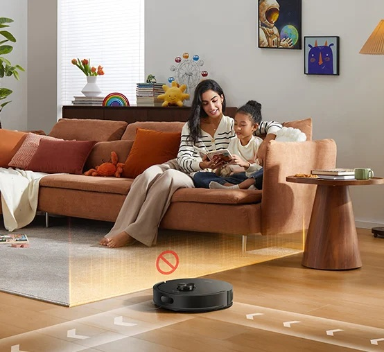 Robot sprzątający czyści podłogę, podczas gdy matka z dzieckiem czyta na kanapie.