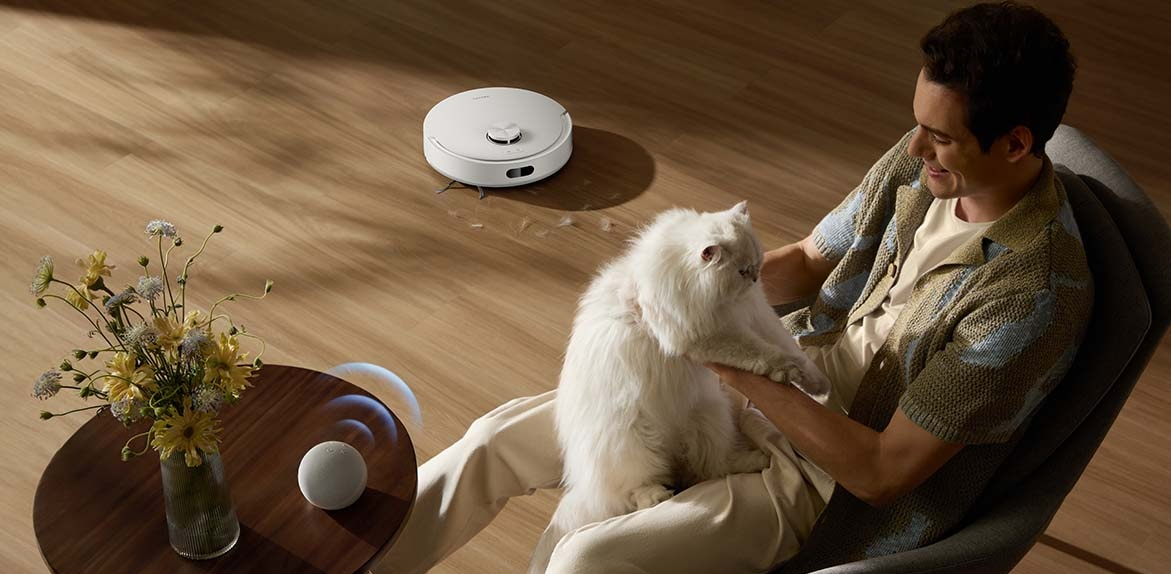 Mężczyzna trzyma białego kota, robot sprzątający czyści podłogę. Kwiaty na stole.