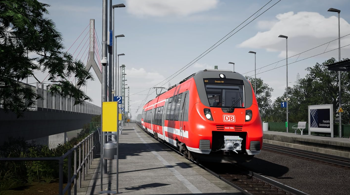 Ein roter Zug steht auf einem Bahnsteig, eine Brücke im Hintergrund.