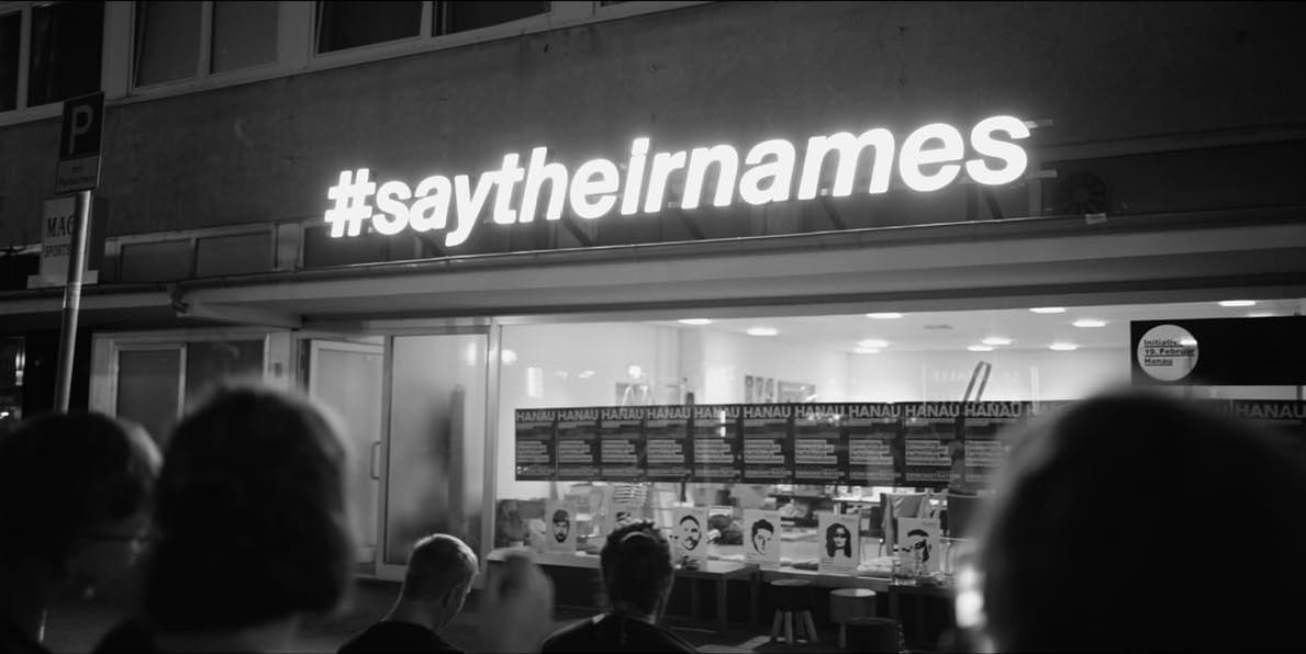 Leuchtende Schrift #saytheirnames über einem Schaufenster mit Fotos und "HANAU"-Aufdruck.