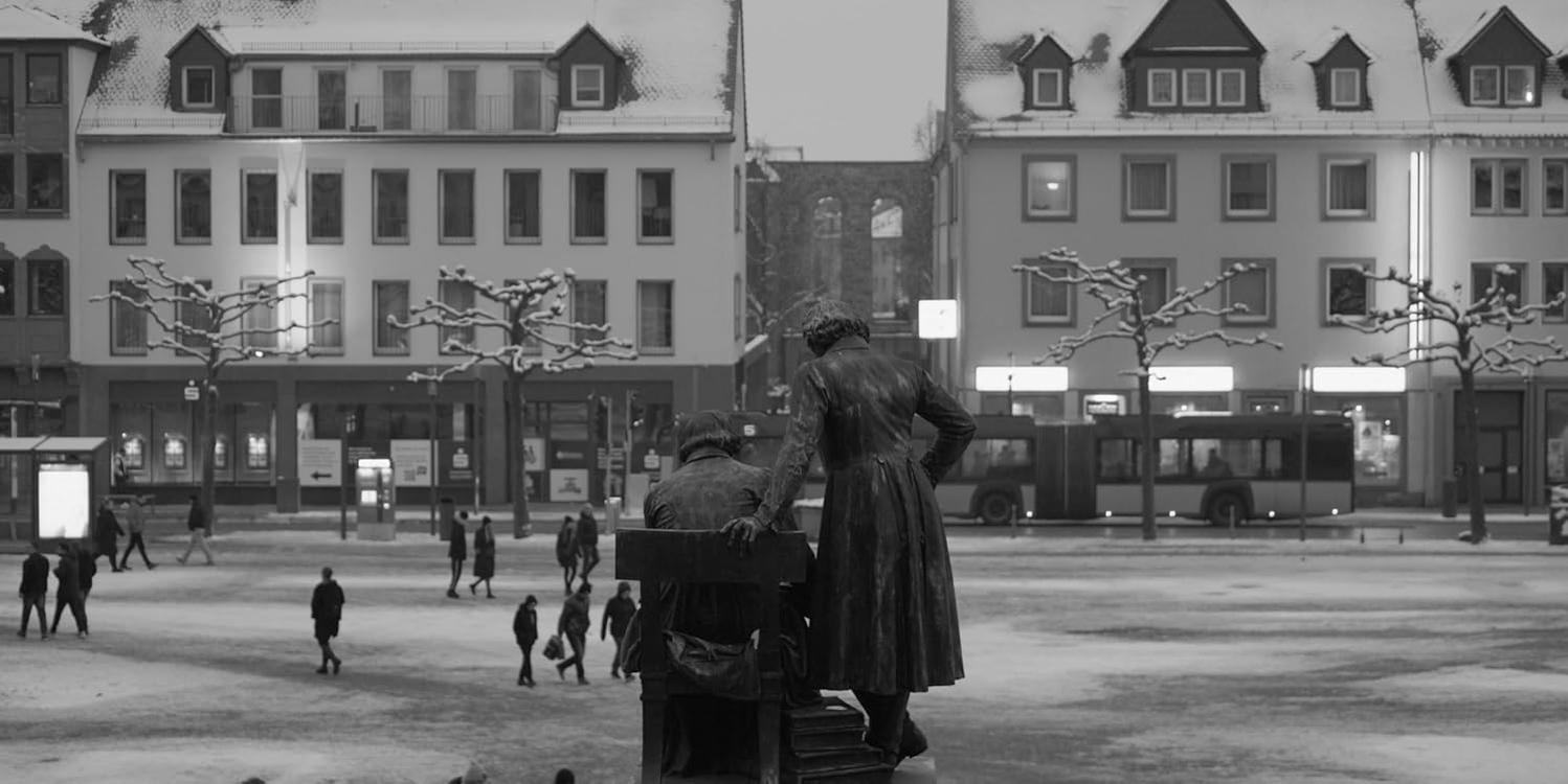 Statuen von zwei Personen auf einem verschneiten Platz vor Gebäuden mit einem Bus im Hintergrund.