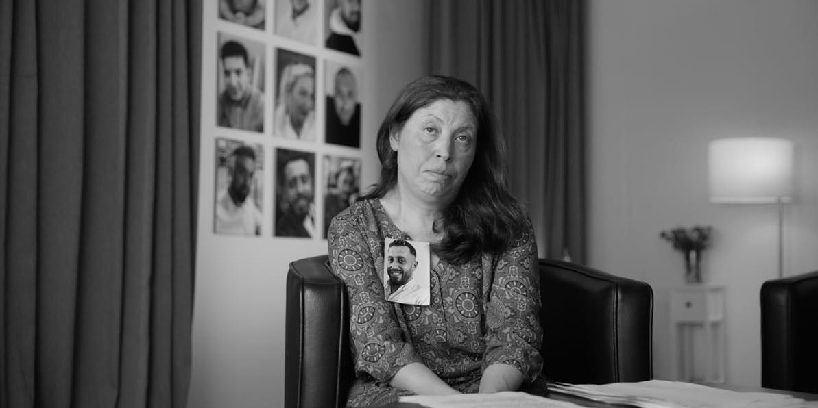 Eine Frau sitzt vor einer Wand mit Fotos und hält ein Foto von einem Mann.