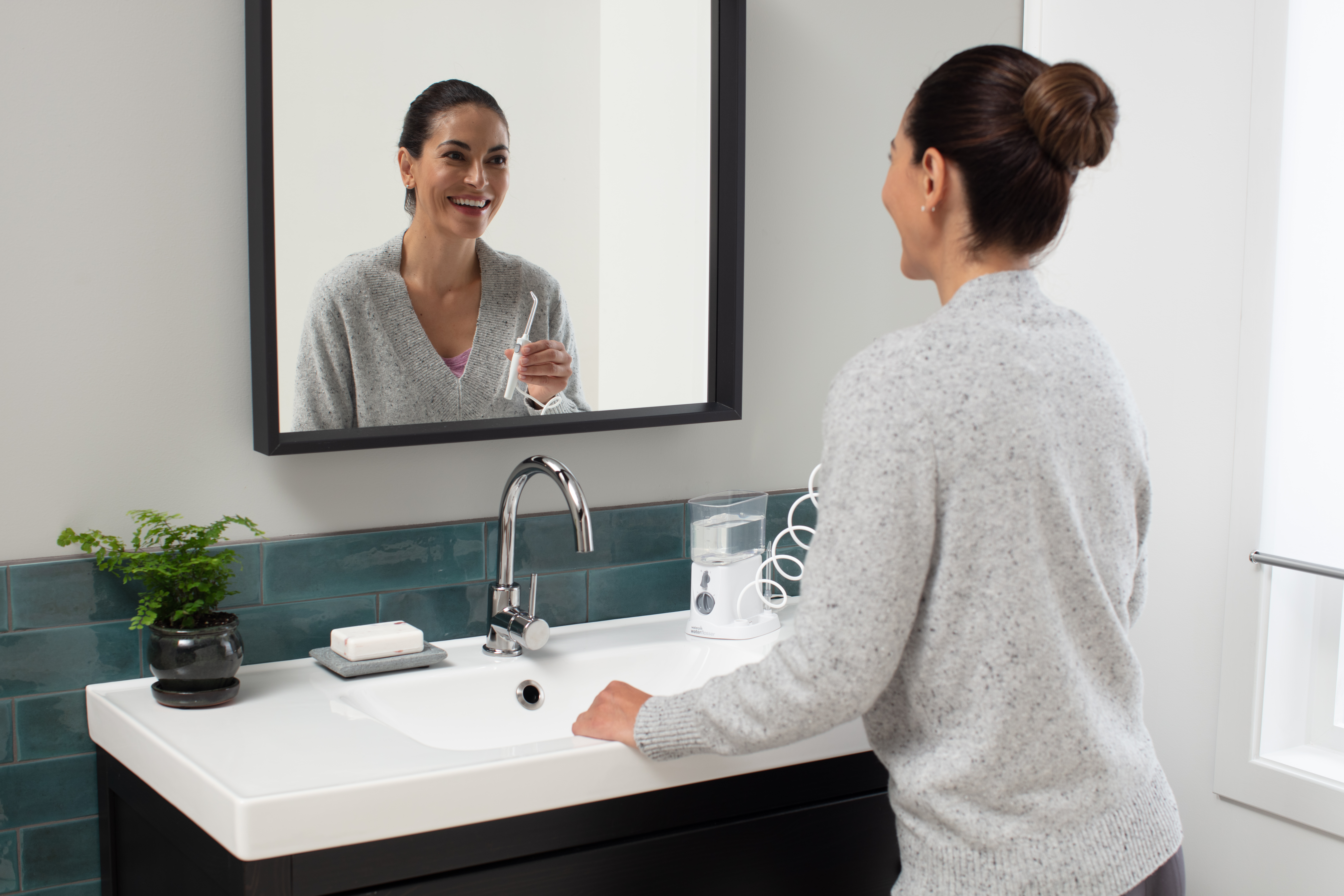 Kobieta w szarym swetrze uśmiecha się w lustrze z szczoteczką. Umywalka.
