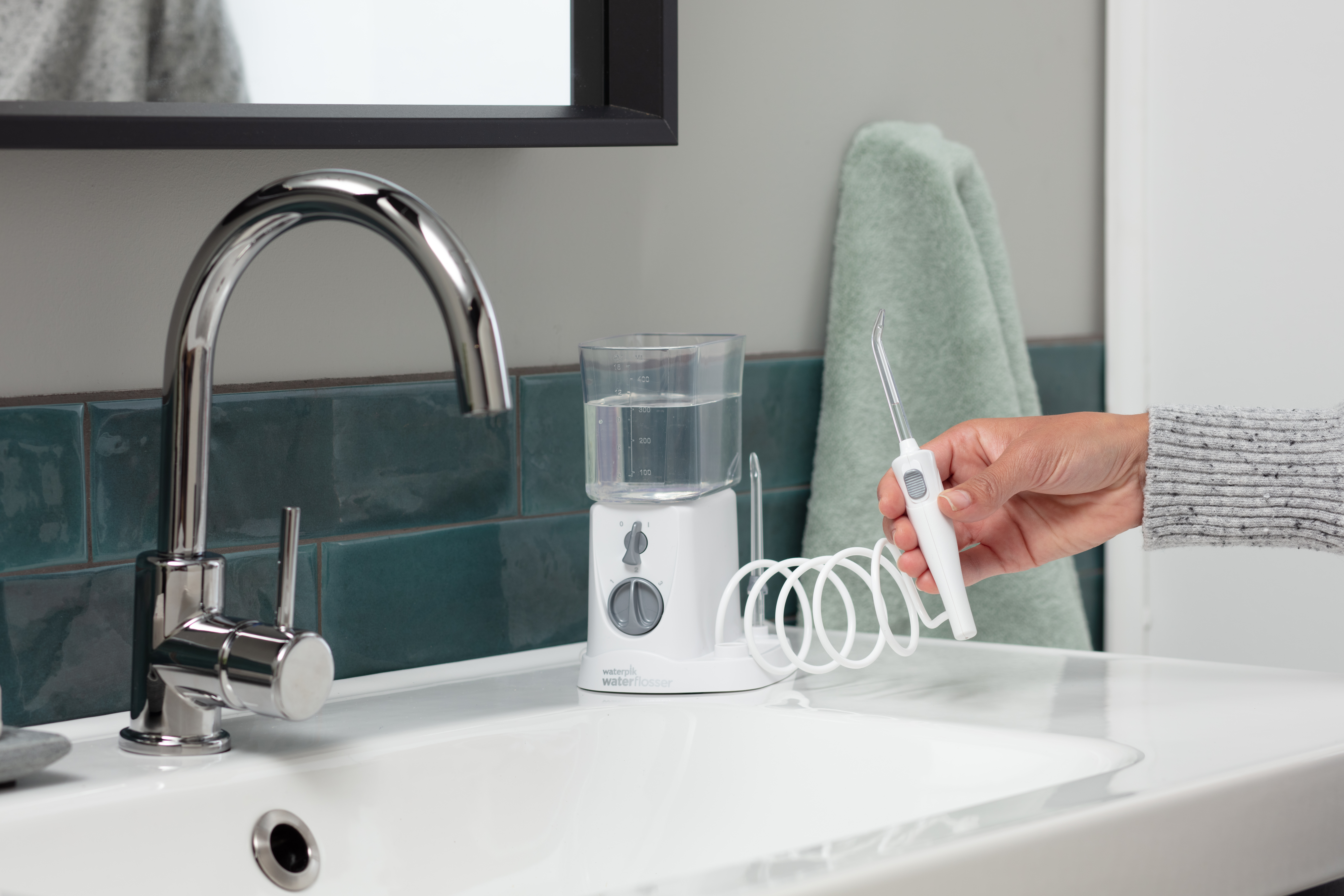 Ręka trzyma białą irygator dentystyczny nad umywalką z kranem i zbiornikiem.