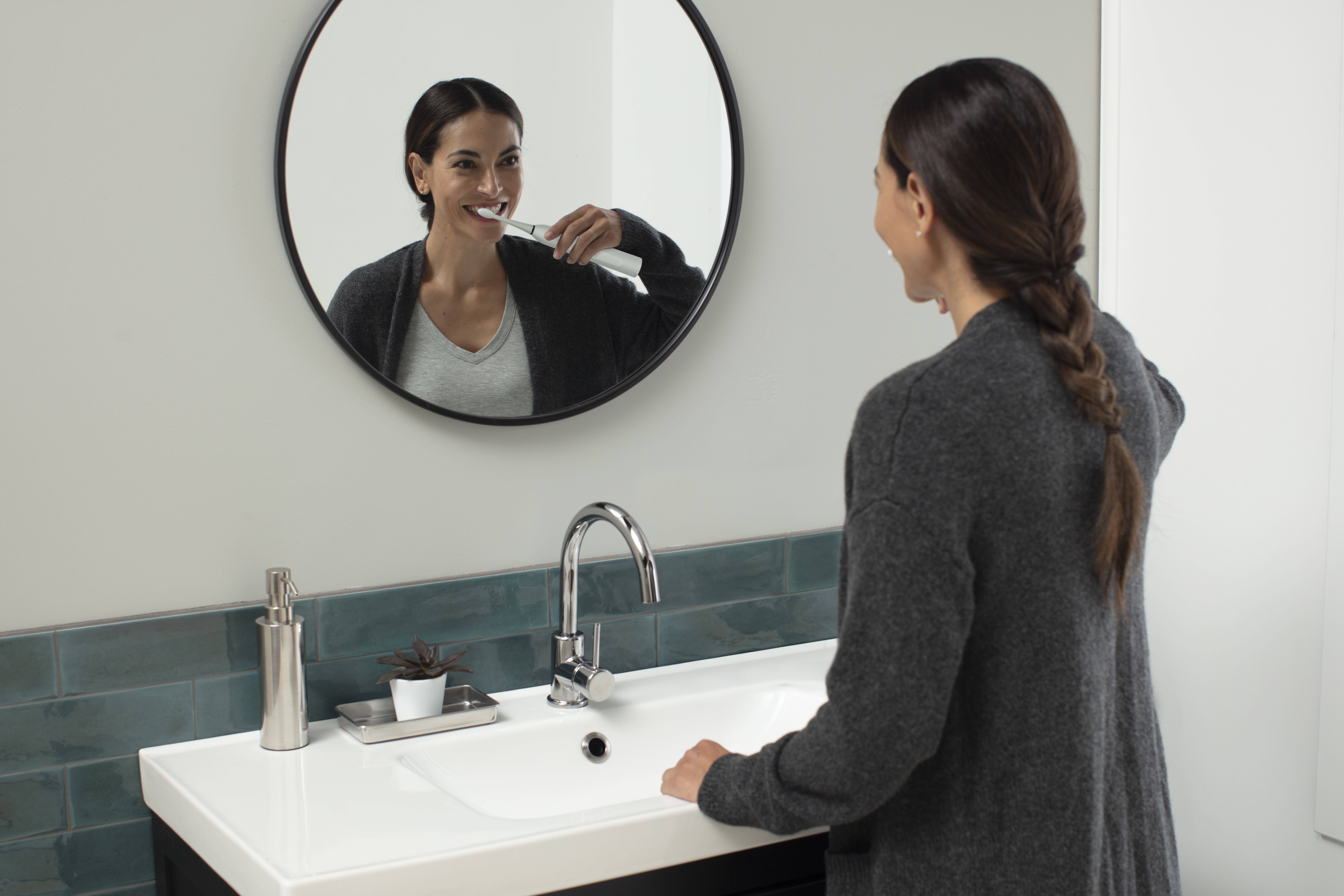 Kobieta myje zęby w łazienkowym lustrze, z umywalką i dozownikiem mydła.