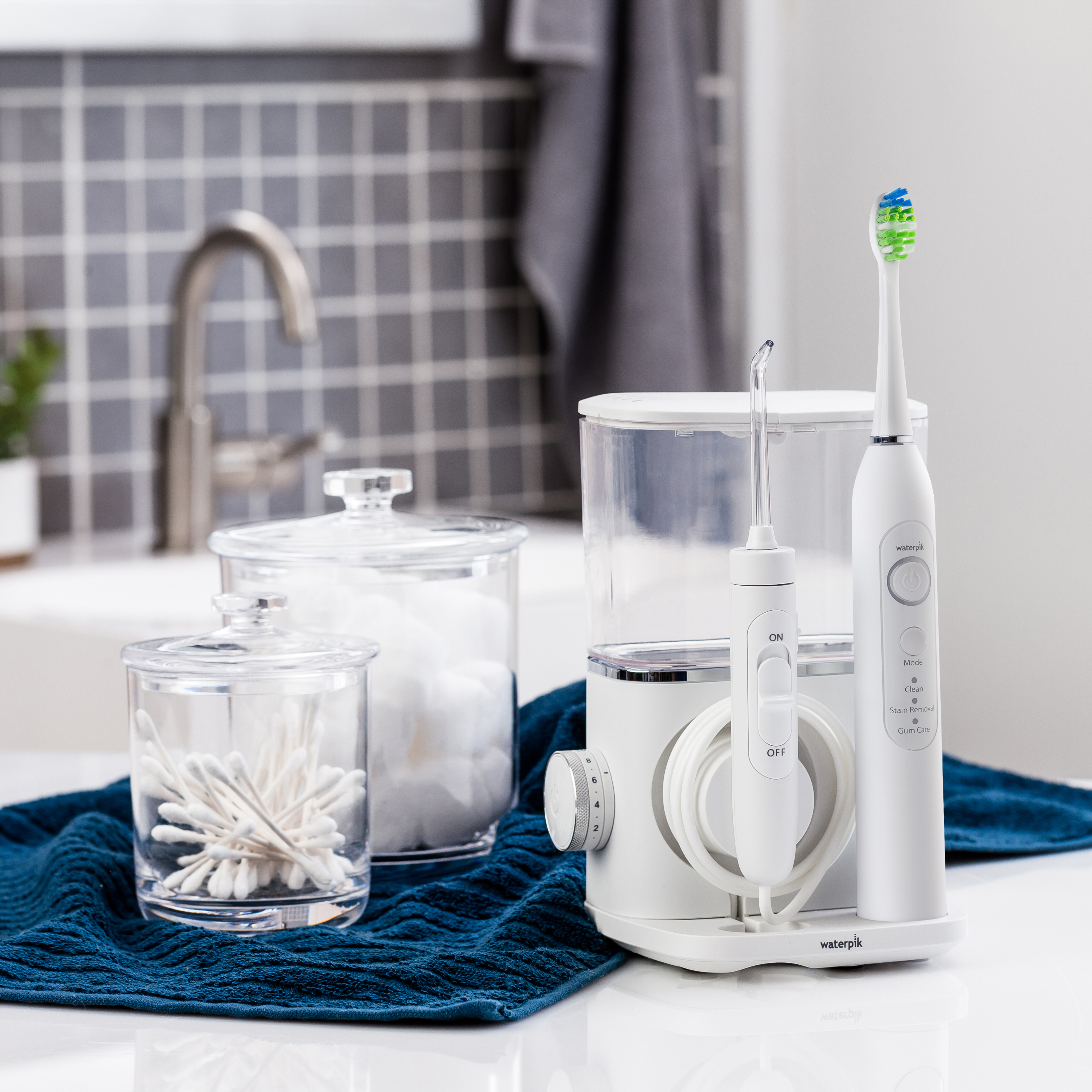 Biały uchwyt na szczoteczkę elektryczną z irygatorem, wacikami i bawełną na niebieskim ręczniku.
