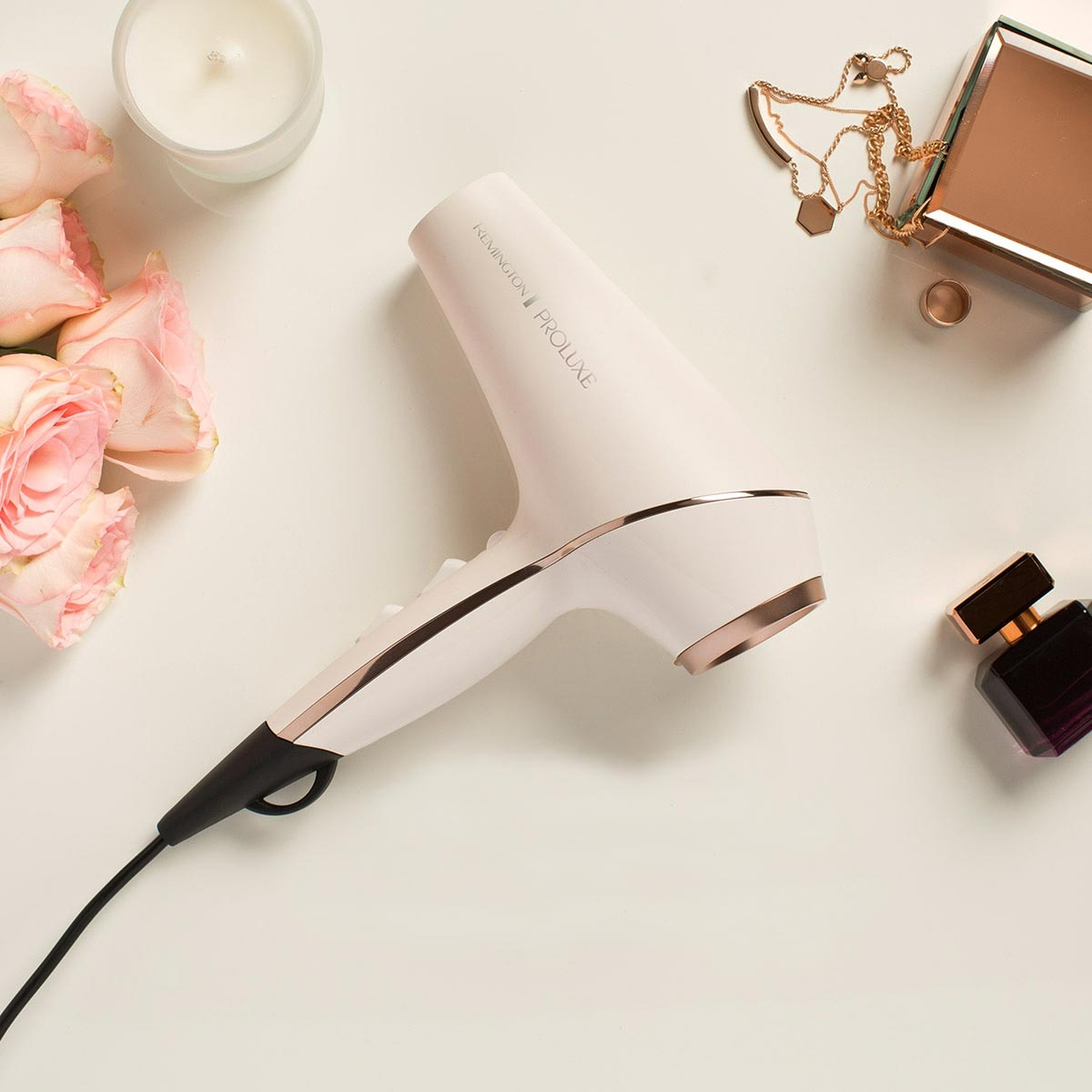 Un asciugacapelli bianco con accenti oro rosa, vicino a rose, candela, gioielli e profumo.