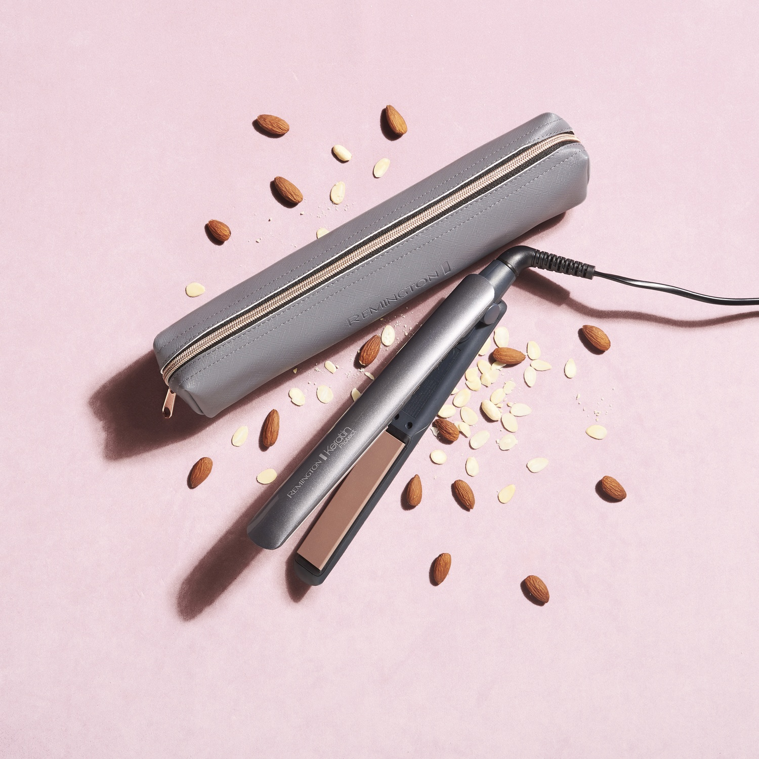 Una piastra per capelli accanto a una custodia grigia, circondata da mandorle su sfondo rosa.