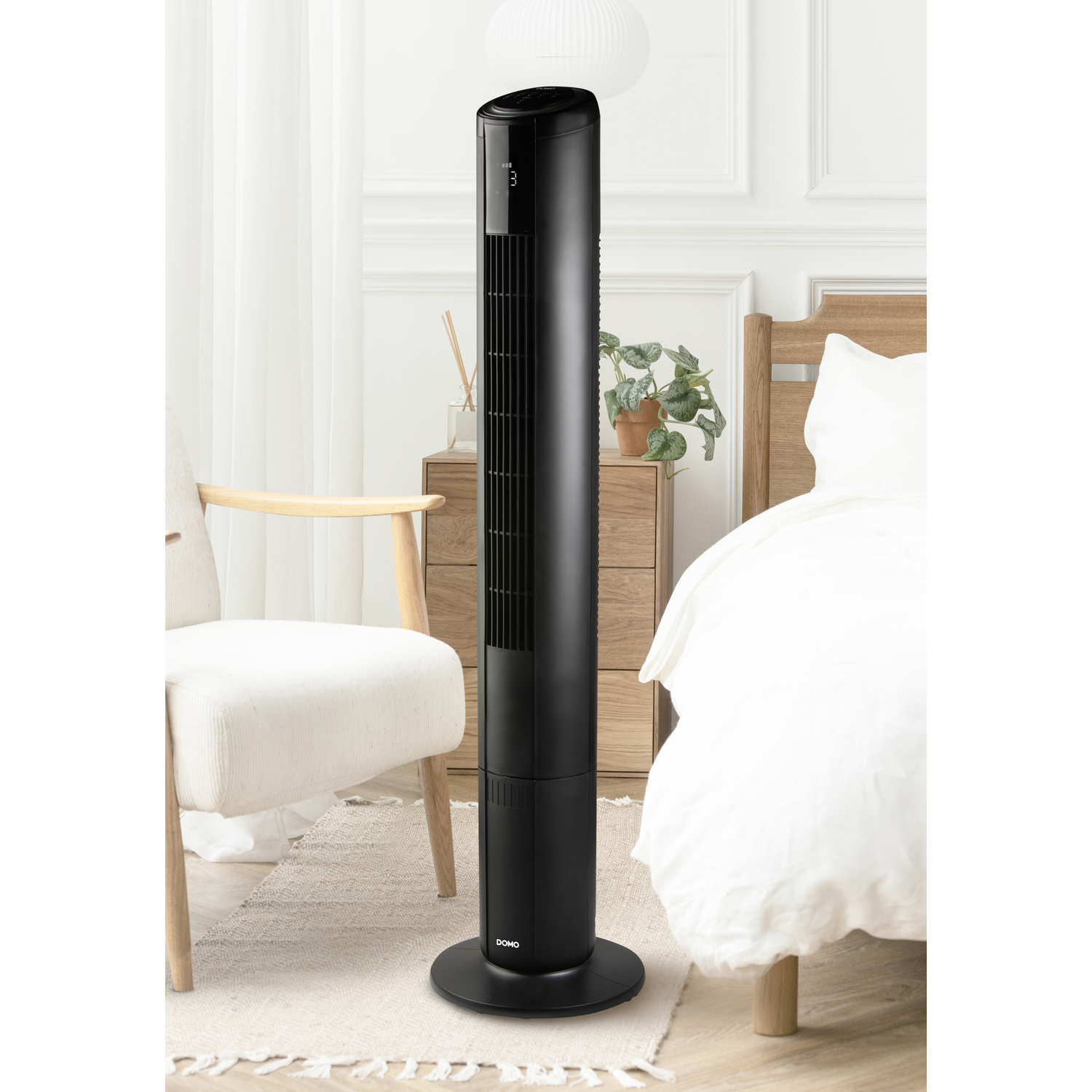 A sleek black tower fan stands beside a bed and an armchair in a room.
