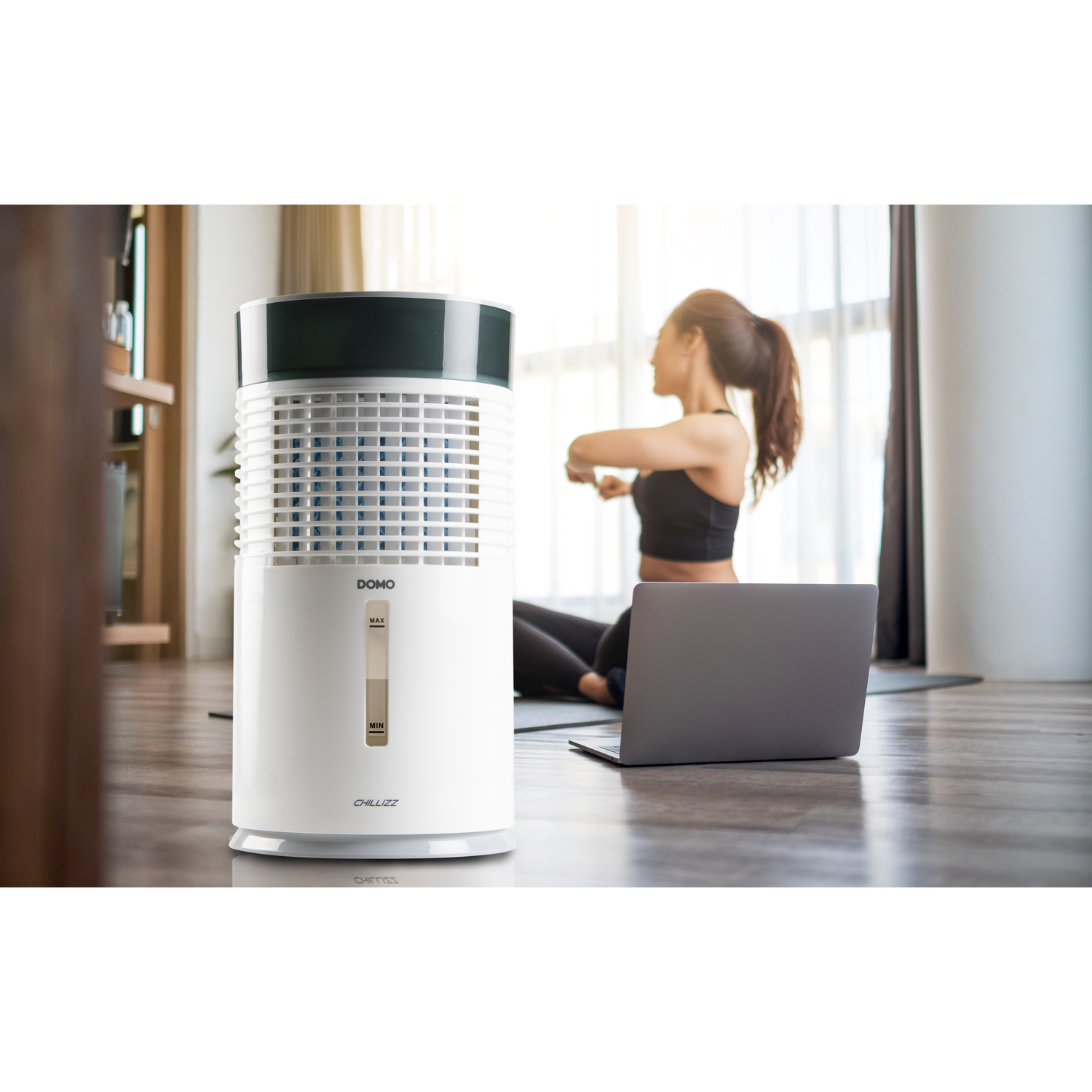A white air cooler sits by a laptop, a woman stretches in the background.