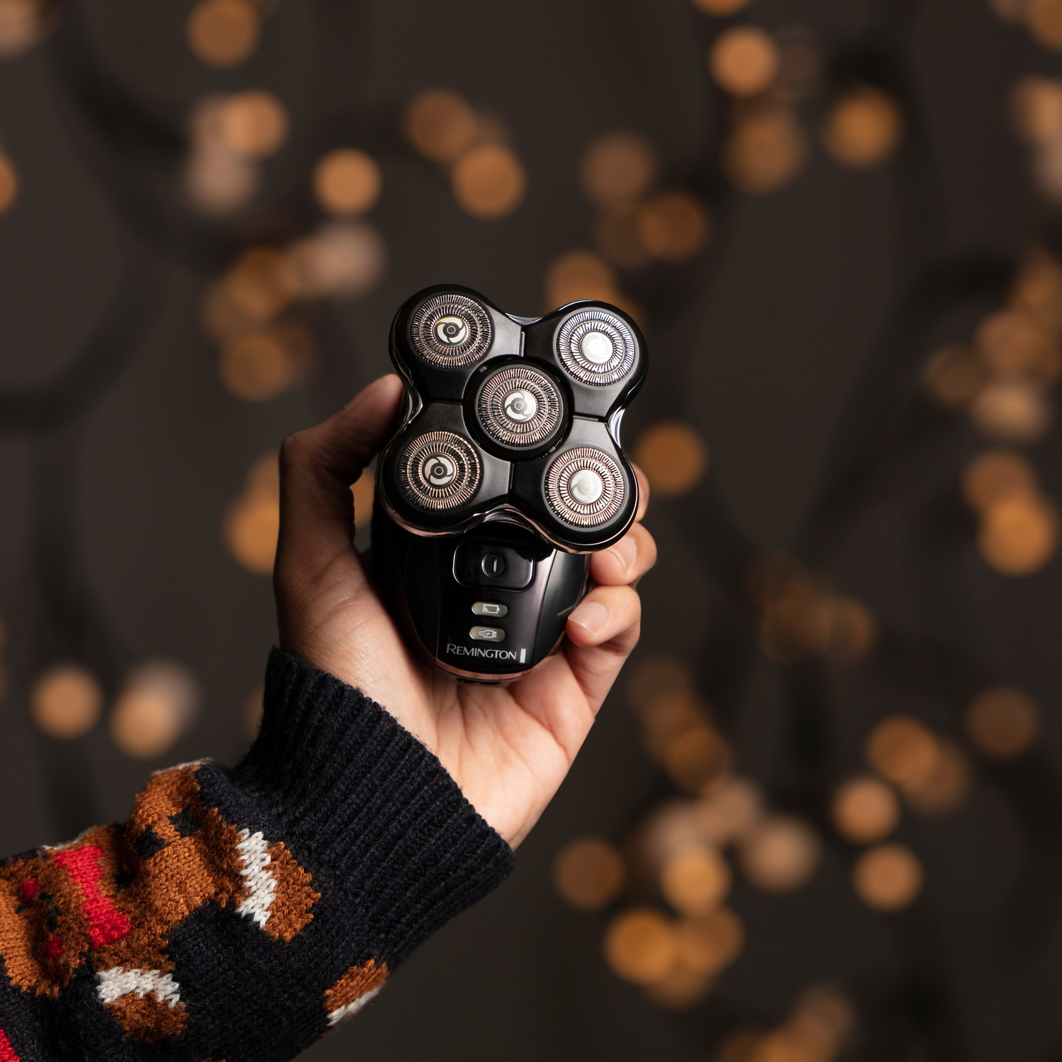 Una mano tiene un rasoio elettrico nero con cinque testine rotanti su sfondo bokeh.