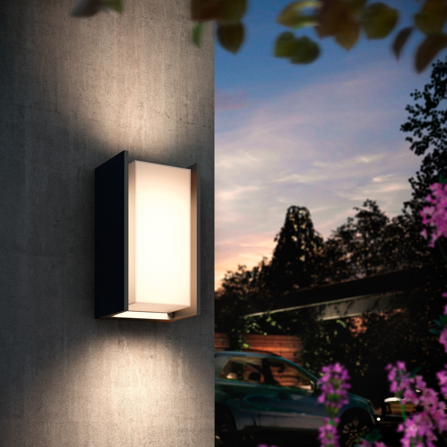 A lit wall lamp cube on a concrete wall. In the background a car and trees at dusk.