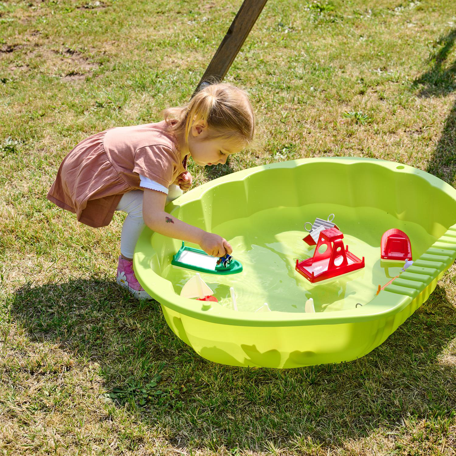 Ein kleines Mädchen spielt mit Spielzeugbooten in einer grünen Sandkastenwanne im Gras.