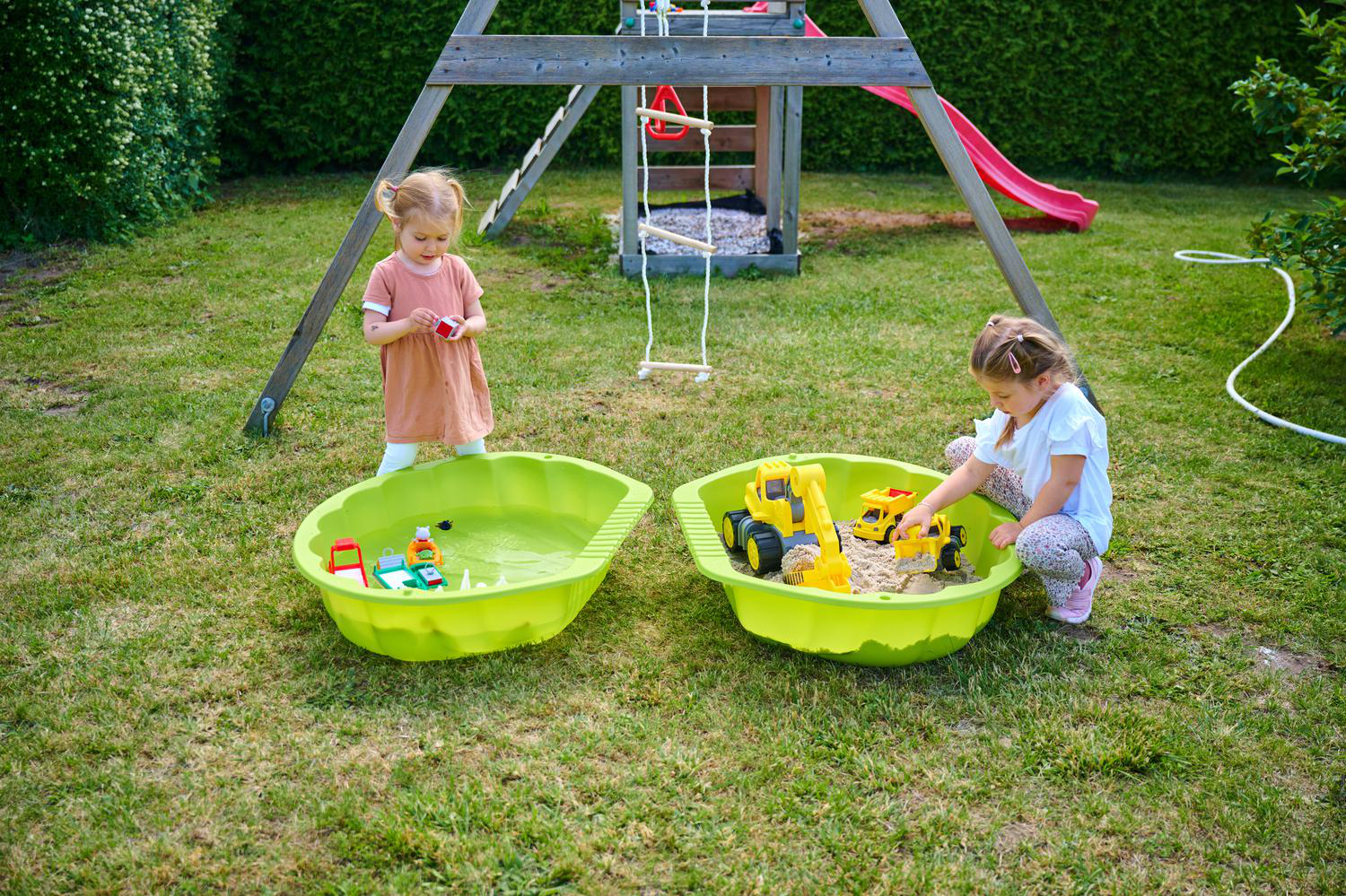 Zwei kleine Mädchen spielen im Garten mit Sand- und Wasserspieltischen neben einem Spielgerüst.