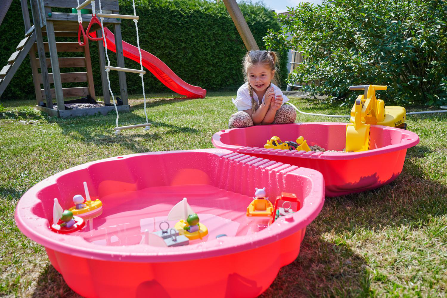 Ein kleines Mädchen sitzt neben zwei rosa Sandkästen mit Spielzeug im Garten.