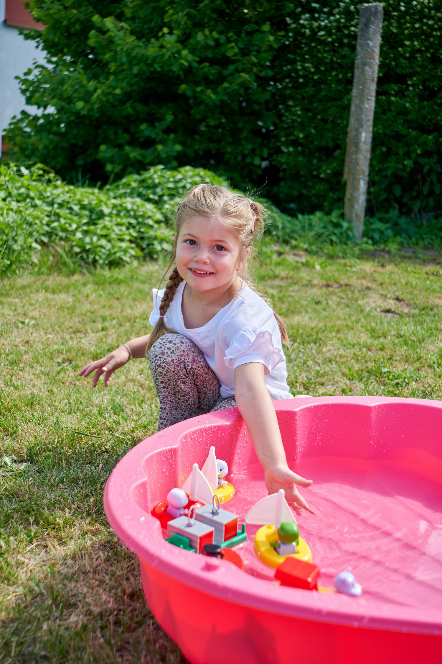 Ein kleines Mädchen spielt mit Spielzeugbooten in einem rosafarbenen Planschbecken im Garten.