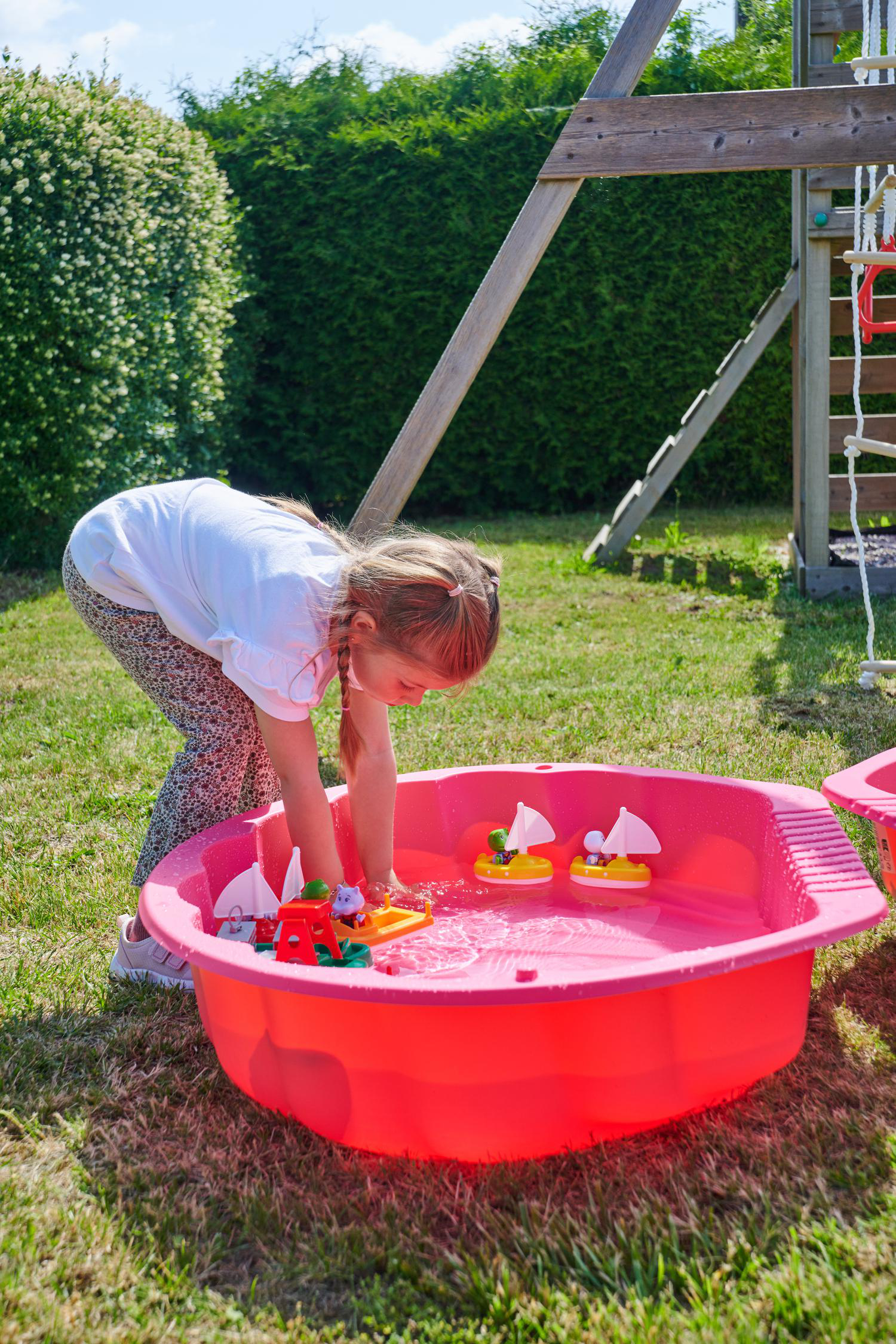 Ein kleines Mädchen spielt mit Spielzeugbooten in einer roten Sandkasten-Wanne im Garten.