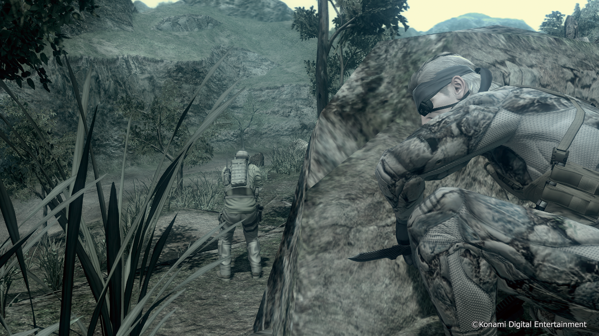 Żołnierz w kamuflażu kuca za skałą, drugi stoi w oddali.