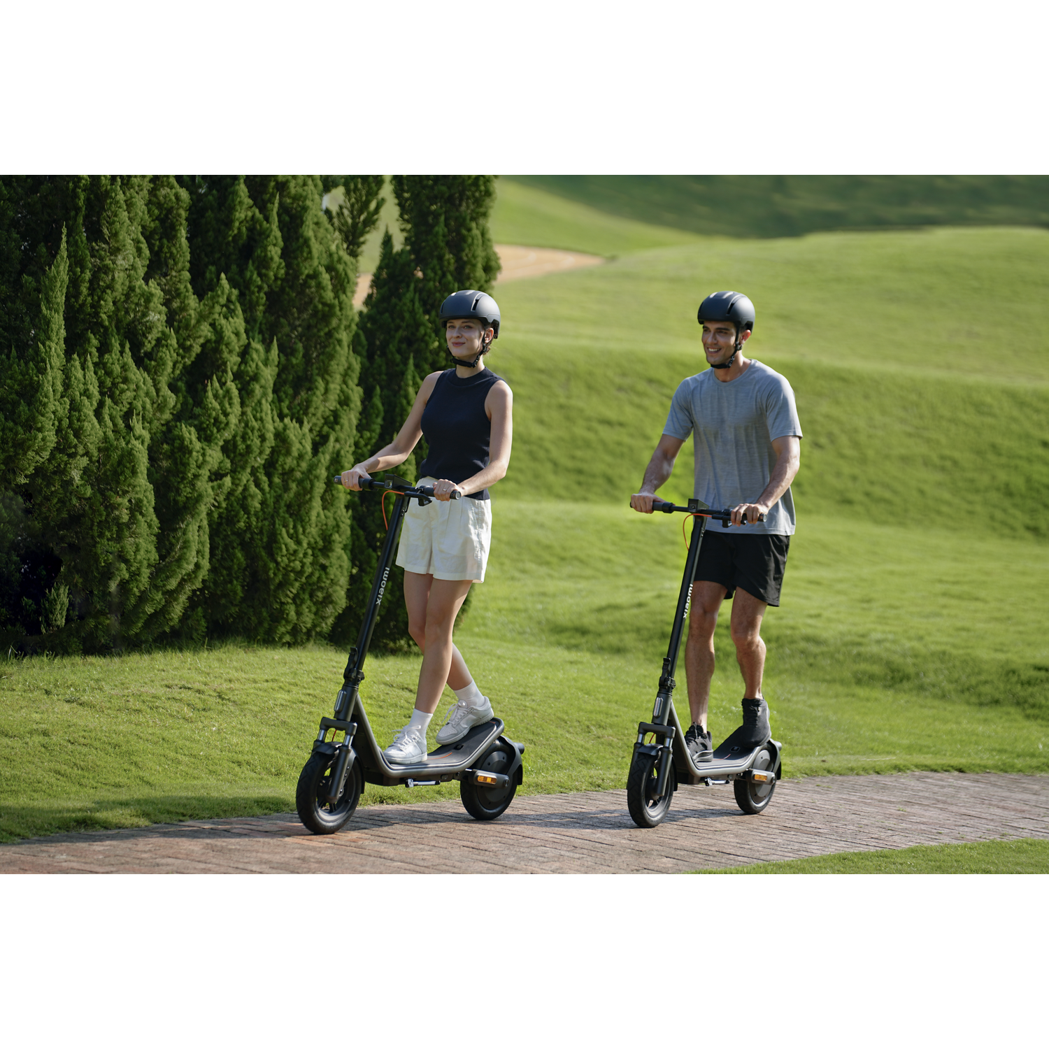Man and woman riding electric scooters on a path with trees and hills in the background.