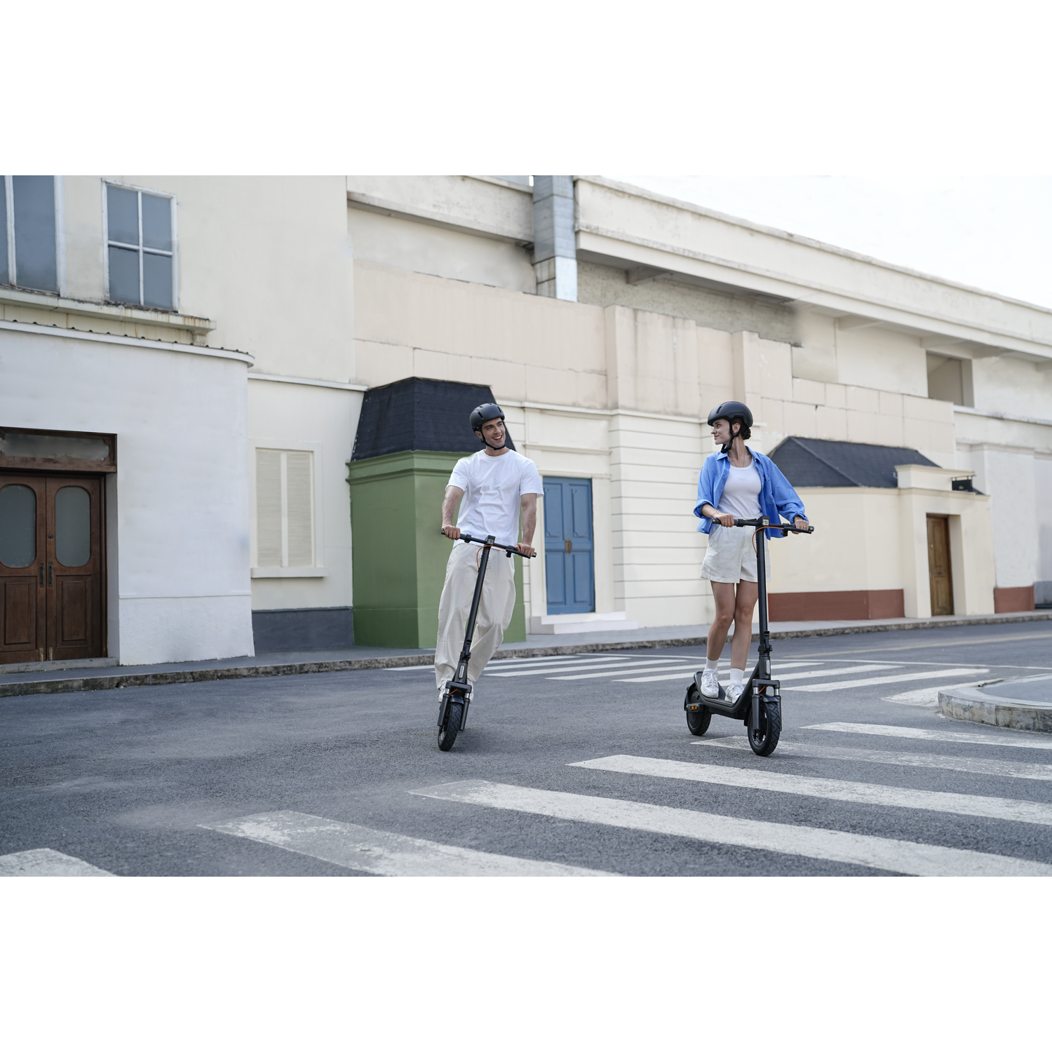 A man and woman ride electric scooters across a crosswalk in a city.