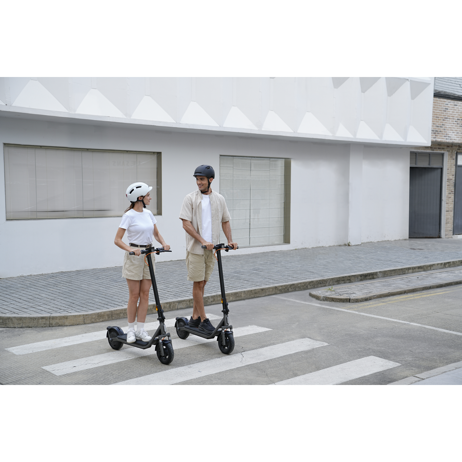 Twee mensen op e-scooters steken een zebrapad over voor een modern gebouw.